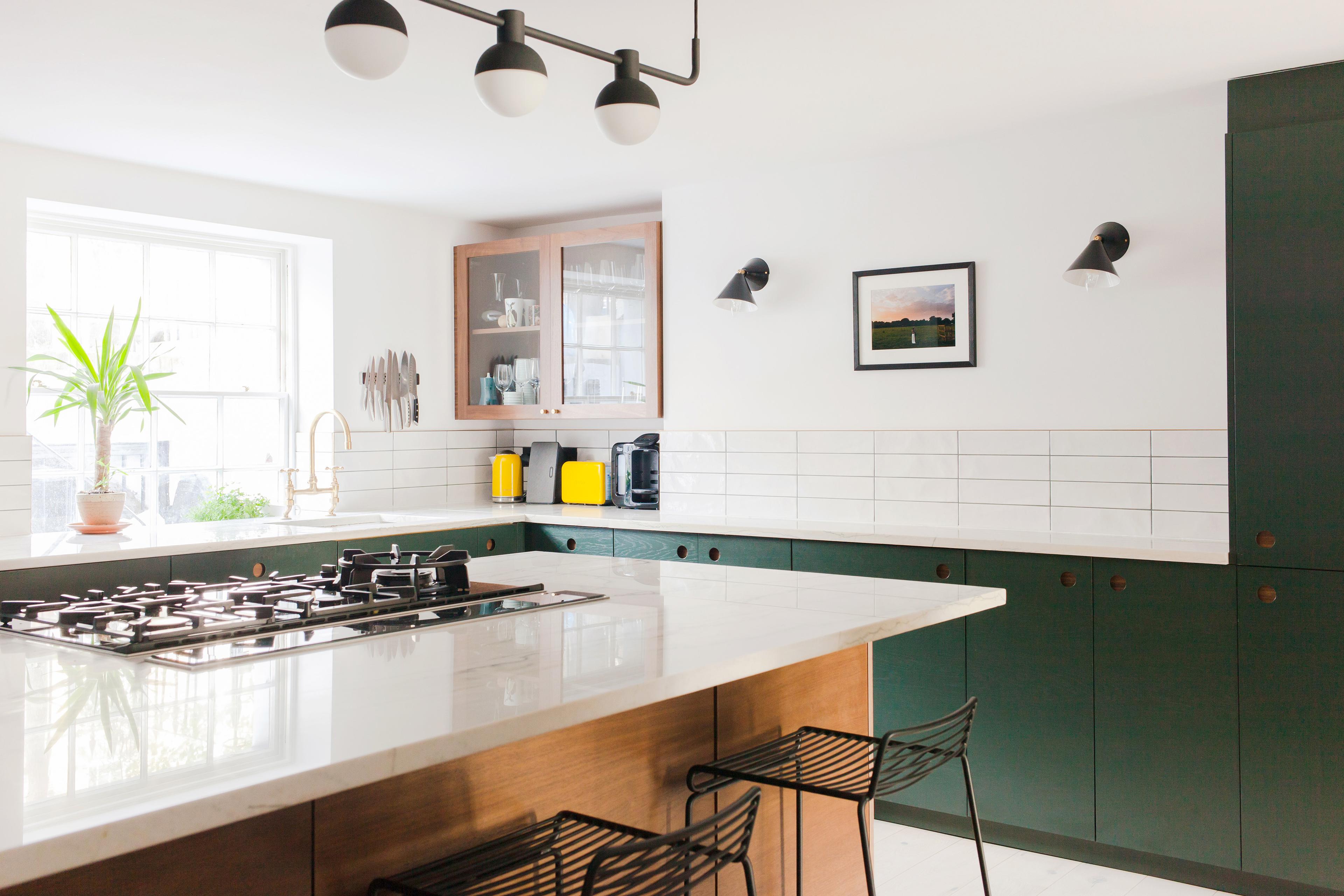 Modern kitchen with white walls and subway tile backsplash. Features a large island with a gas cooktop, wooden and green cabinets, a window with a plant, wall-mounted lighting, and a framed picture. Two black stools are placed at the island.