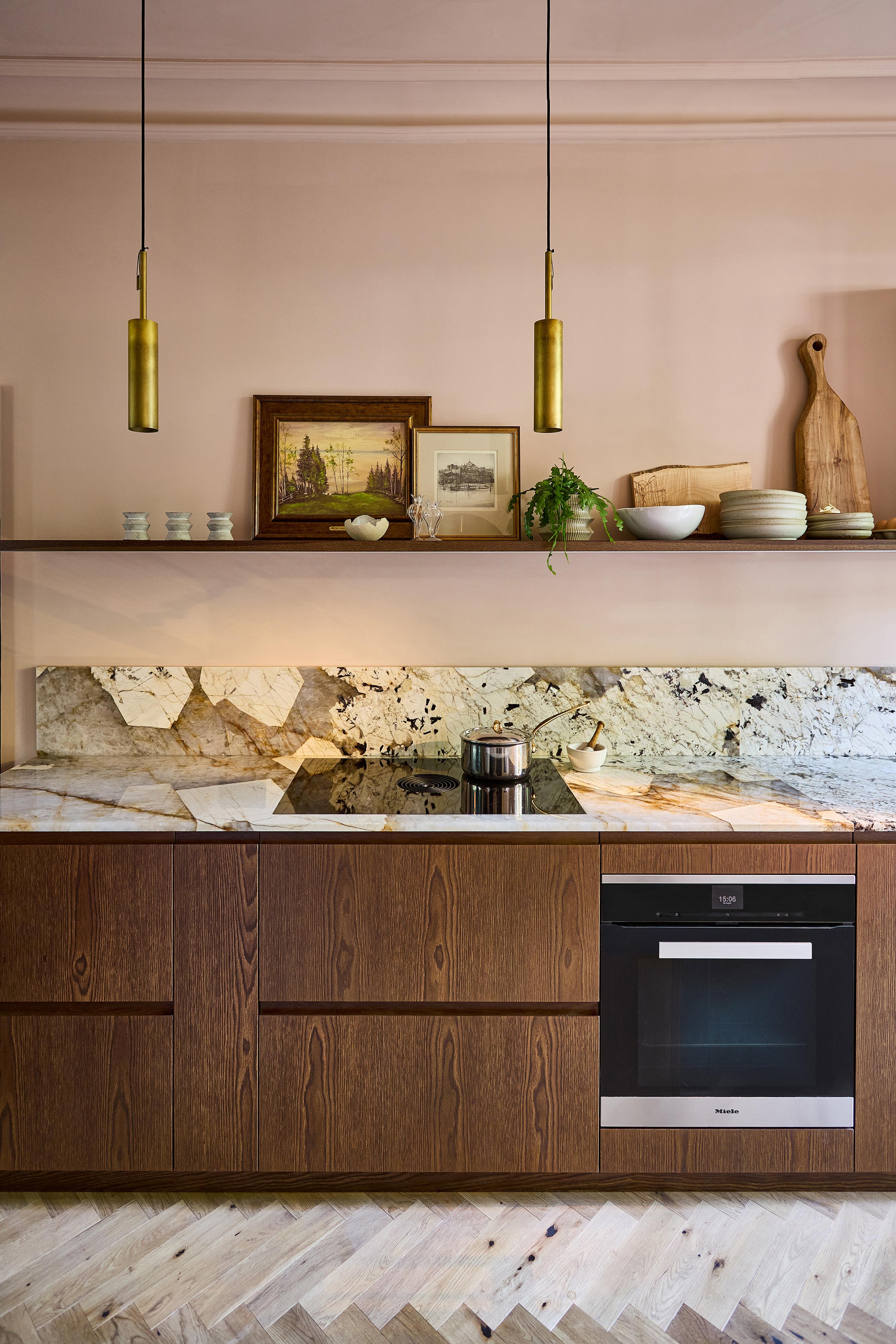 A modern kitchen with wooden cabinets, marble backsplash, a built-in oven, cookware on the stove, open shelves holding plates, bowls, framed art, and decorative items, lit by two gold pendant lights.