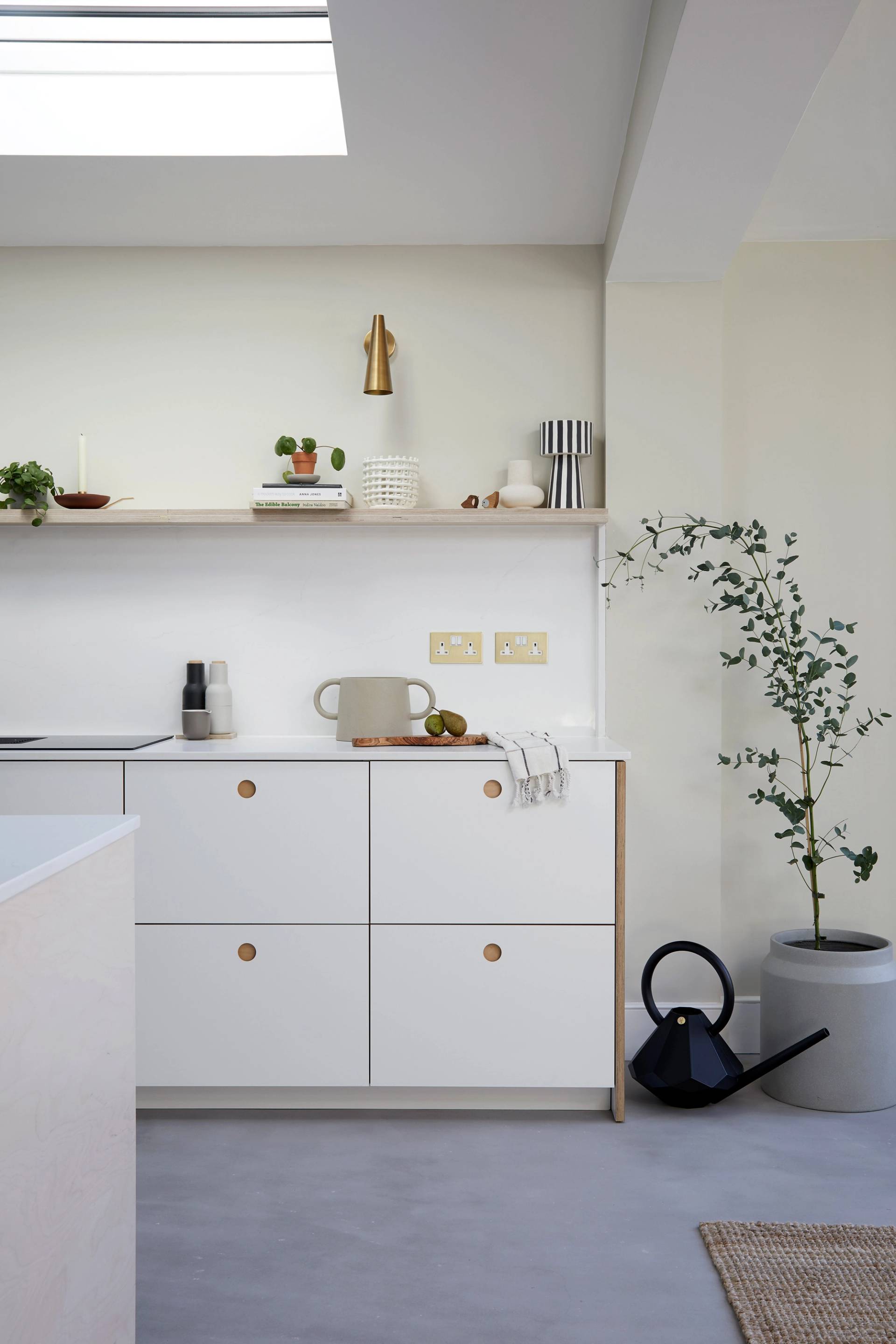 Minimalist kitchen with white cabinets featuring round wooden knobs, open shelving with plants, ceramics, and a brass accent piece. A potted plant and black watering can on the floor. Light streaming through a skylight. Neutrally toned decor.