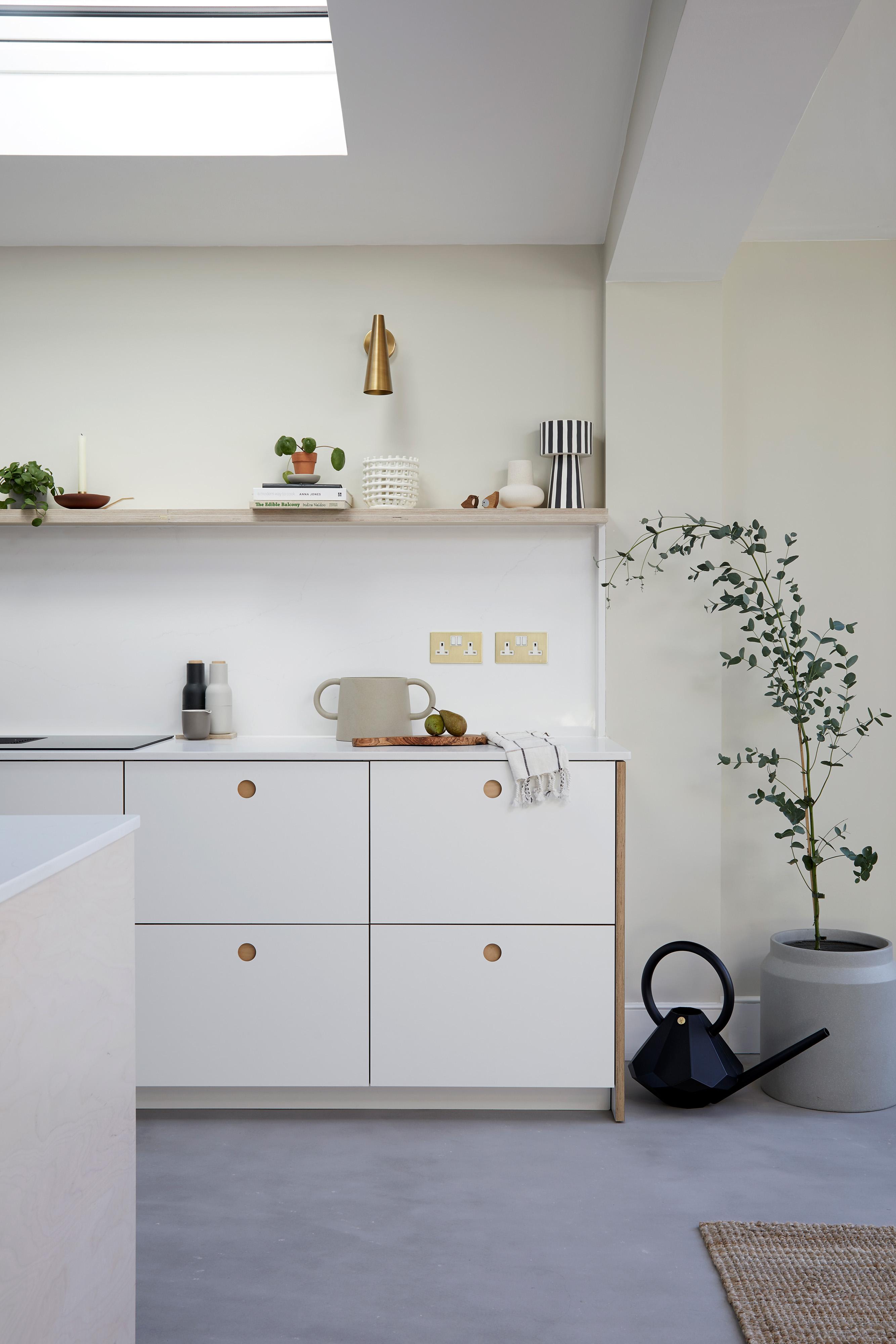 Minimalist kitchen with white cabinets featuring round wooden knobs, open shelving with plants, ceramics, and a brass accent piece. A potted plant and black watering can on the floor. Light streaming through a skylight. Neutrally toned decor.