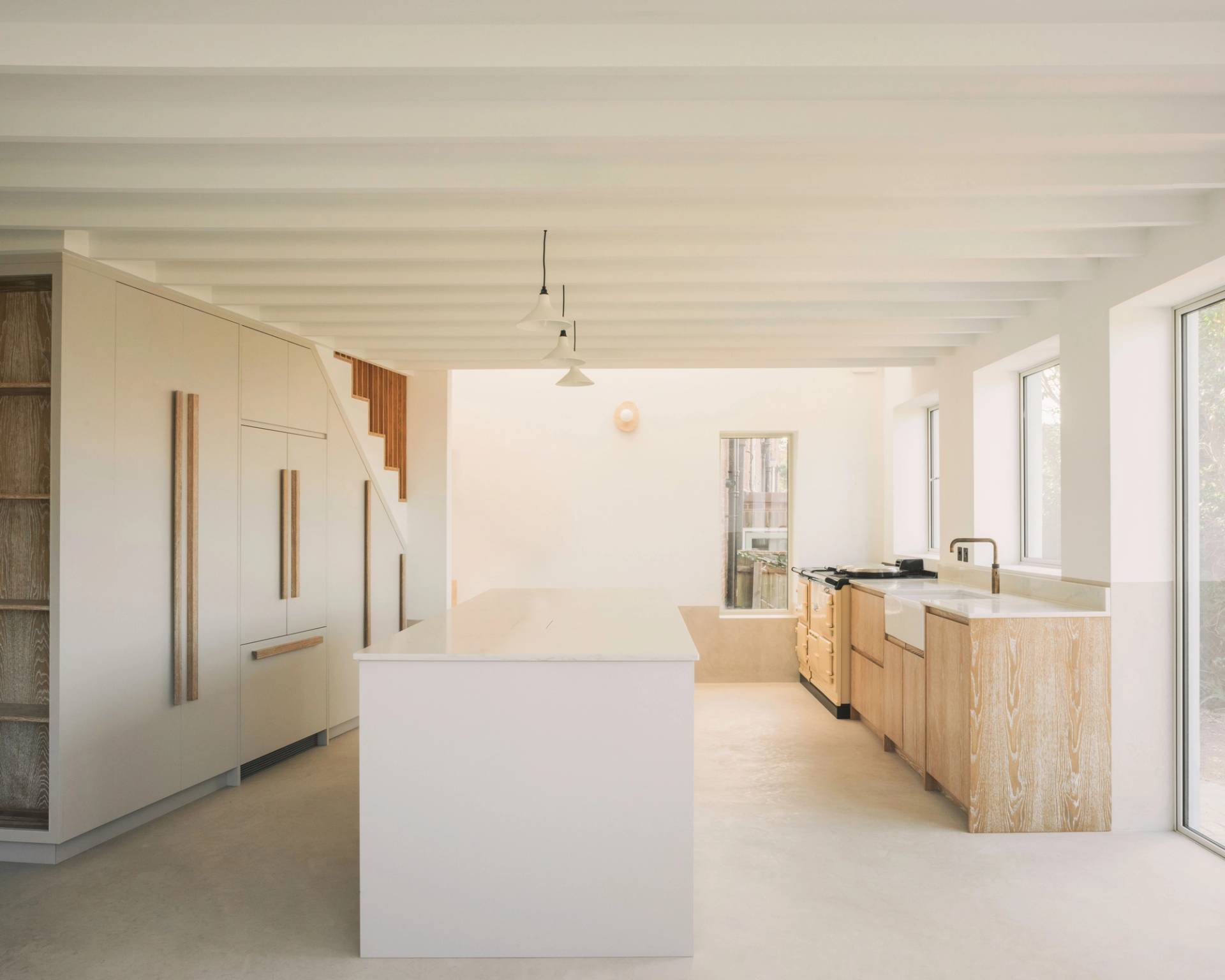 Minimalist kitchen with light wood cabinets, a white island countertop, built-in appliances, pendant lights, and large windows letting in natural light. Exposed ceiling beams and neutral tones create an airy, modern feel.