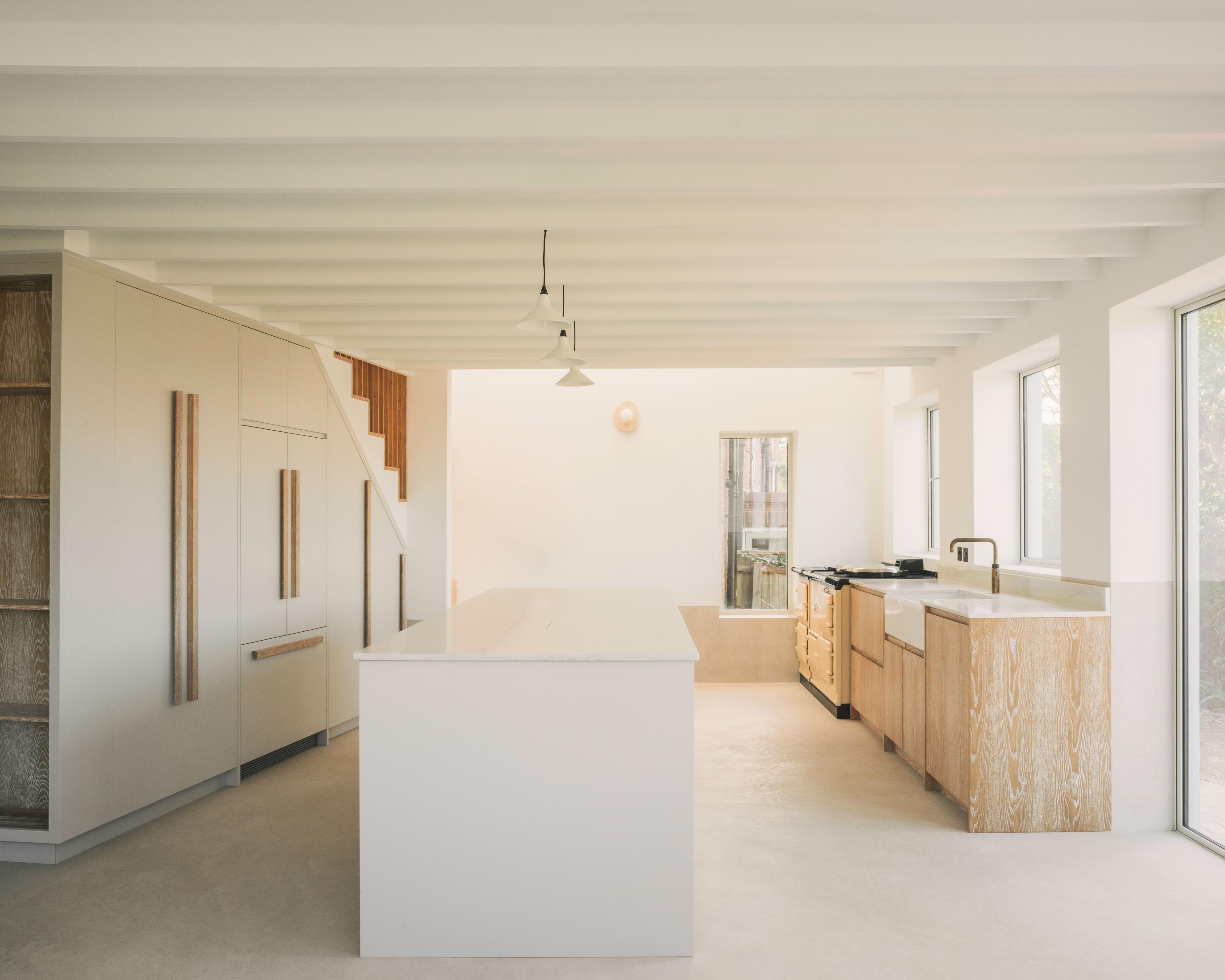 Minimalist kitchen with light wood cabinets, a white island countertop, built-in appliances, pendant lights, and large windows letting in natural light. Exposed ceiling beams and neutral tones create an airy, modern feel.