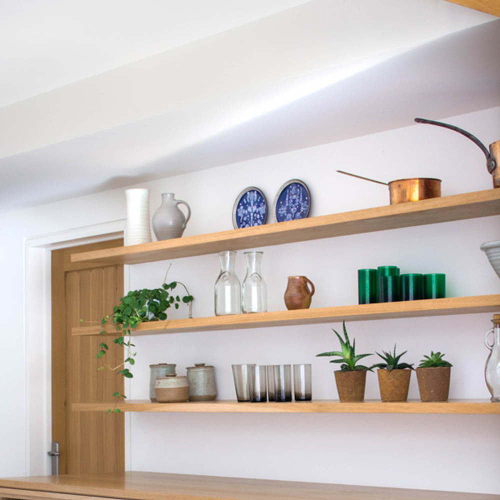 Open wooden shelves with various items, including ceramic plates, glass bottles, metal cups, green glass tumblers, copper cookware, a white vase, potted plants, and a trailing vine, against a white wall.