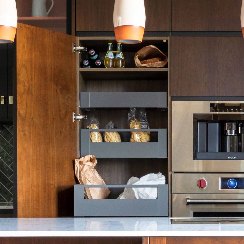A modern kitchen with a wooden cabinet open, revealing three shelves stocked with bottled drinks, pasta in bags, and a paper bag. Adjacent is a built-in stainless steel oven and coffee machine. Pendant lights hang from the ceiling