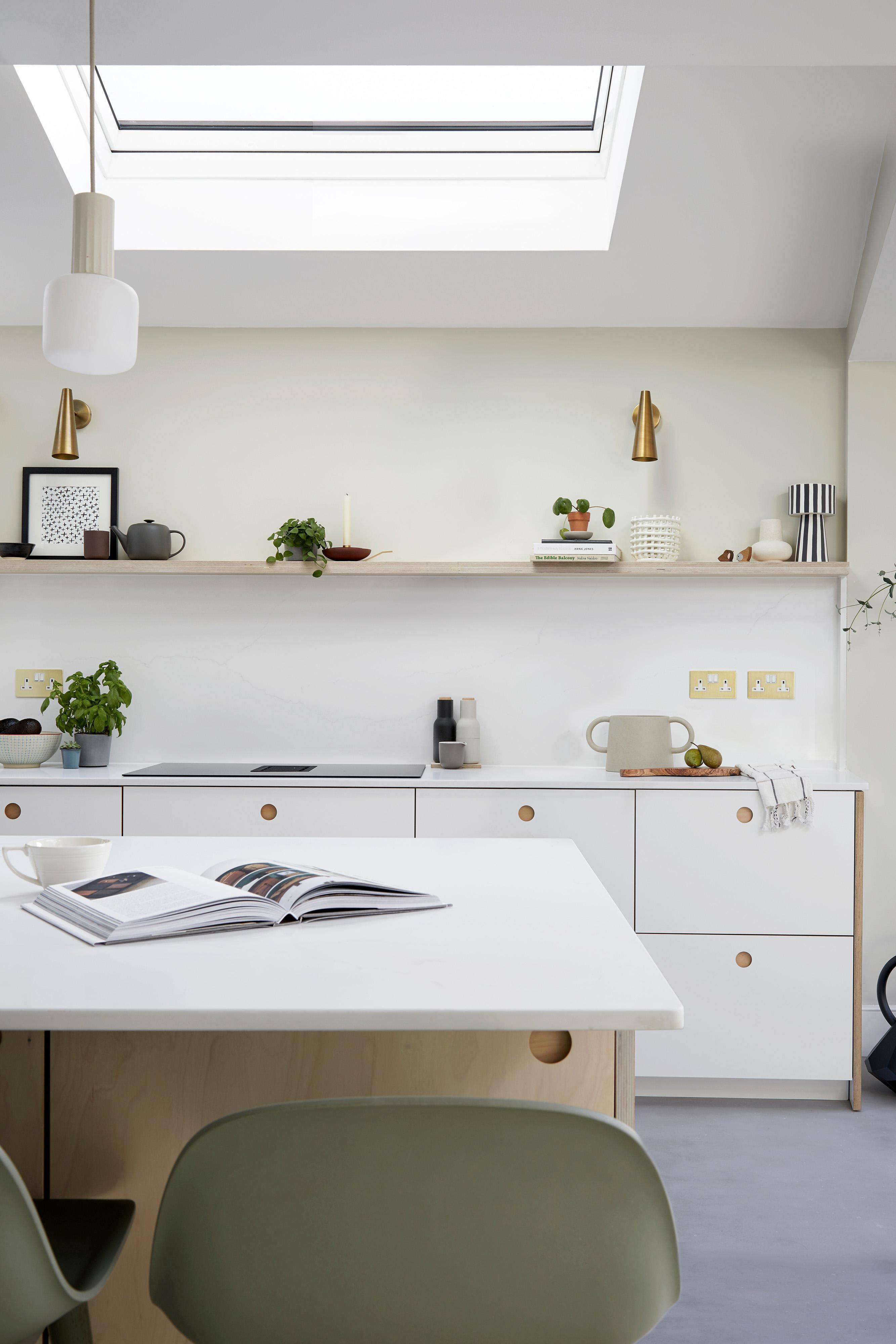 Modern kitchen interior with white cabinetry and a minimalist aesthetic. An open book is placed on a central island countertop. Shelves display small plants, kitchenware, and decor items. A skylight above illuminates the room with natural light.