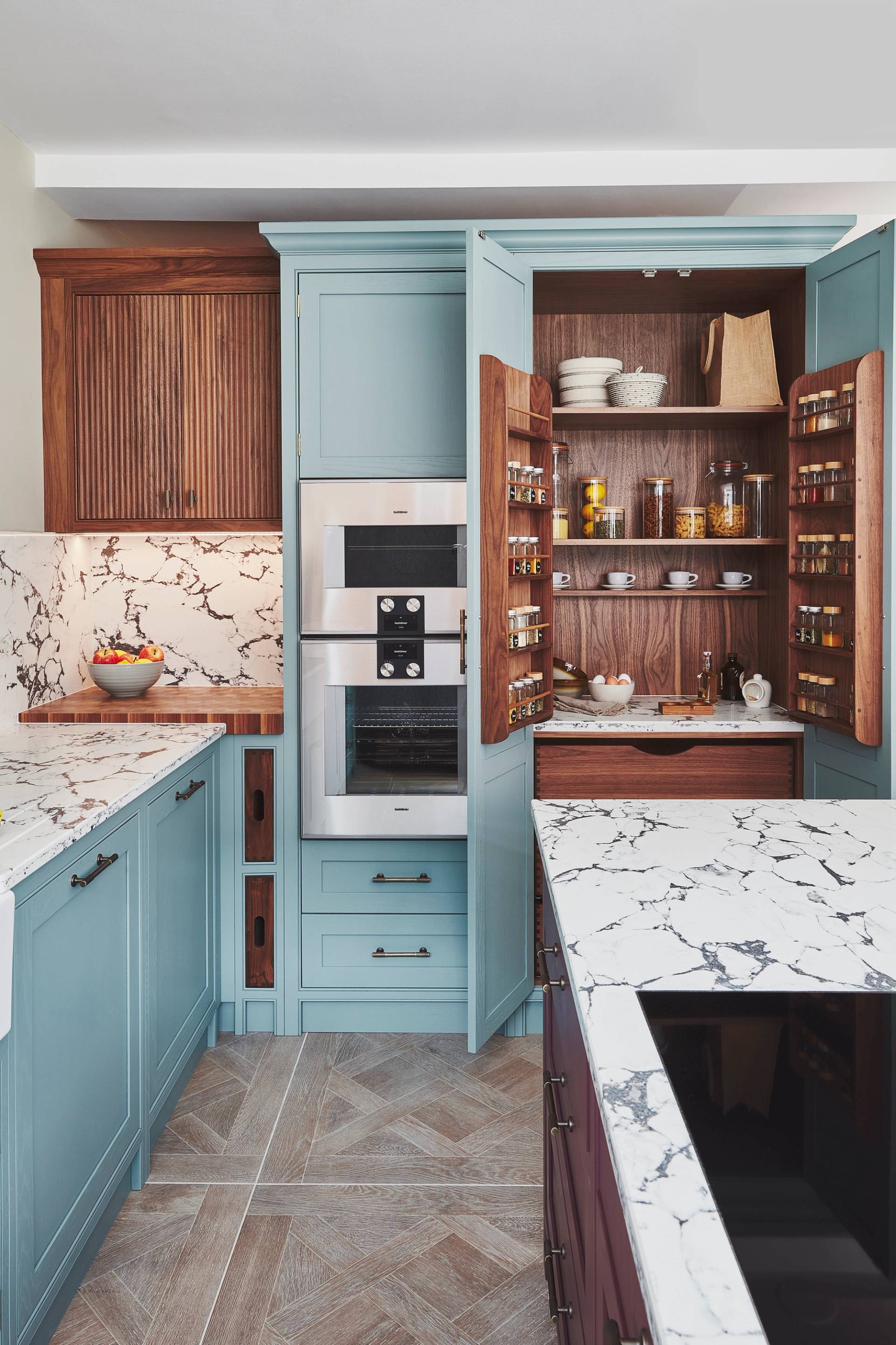 A modern kitchen with light blue cabinets and marble countertops. The pantry cabinet doors are open, revealing shelves stocked with spices and kitchenware. An oven is integrated into the cabinetry. A bowl of fruit is on the counter.