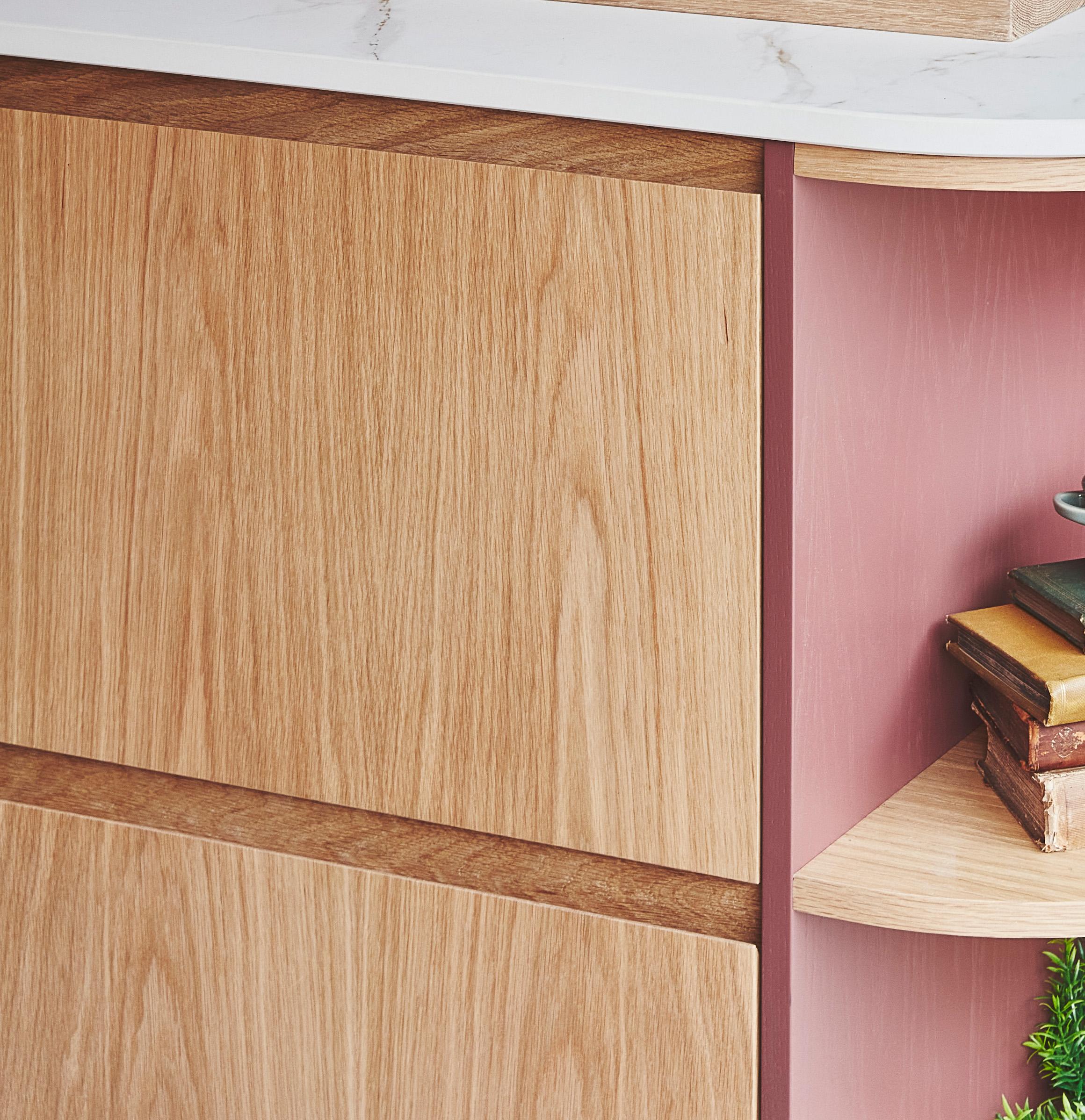 Close-up of a wooden cabinet with a light wood finish. To the right, there is a small pink shelf with rounded edges, holding a stack of books and a small green plant. The countertop above is white with subtle gray veining.