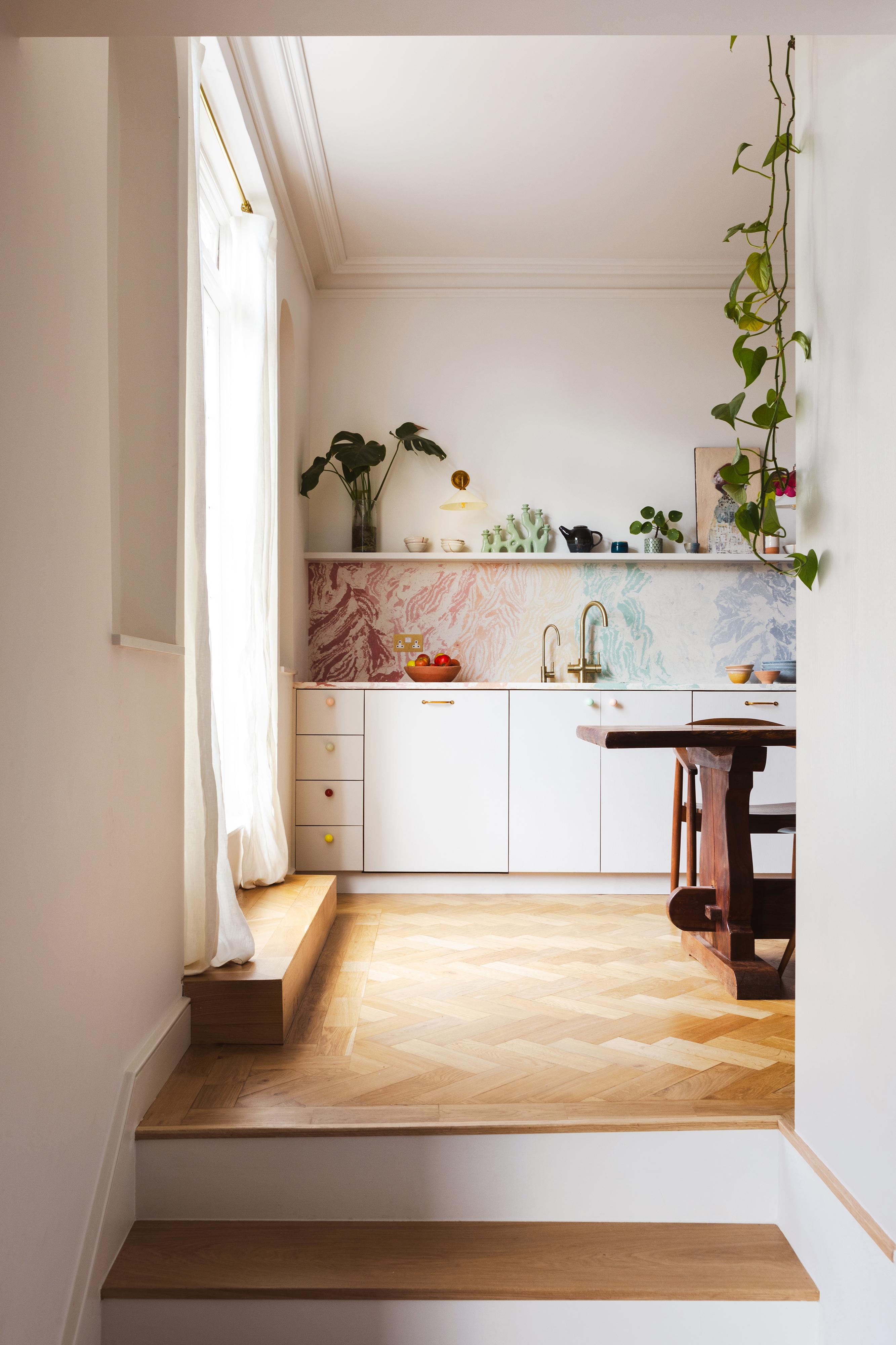 A small kitchen with bright natural light, featuring white cabinets and a colorful marble-patterned backsplash. Wooden countertops hold potted plants. A large plant hangs from above, and a wooden table with chairs sits to the right. Light parquet flooring.