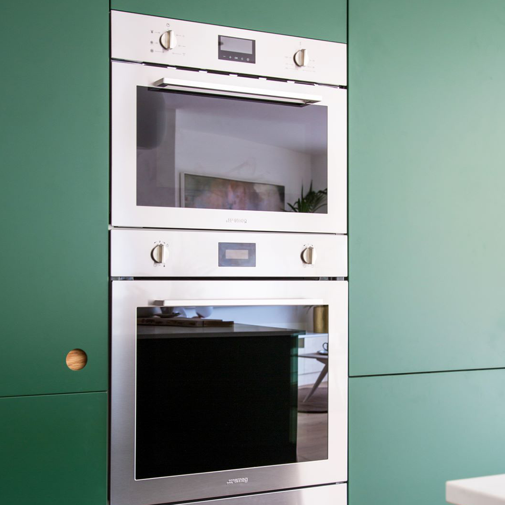 Double built-in stainless steel ovens are installed within a sleek green kitchen cabinet. The ovens have digital displays and control knobs, and a small round wooden handle is visible on the left cabinet door.