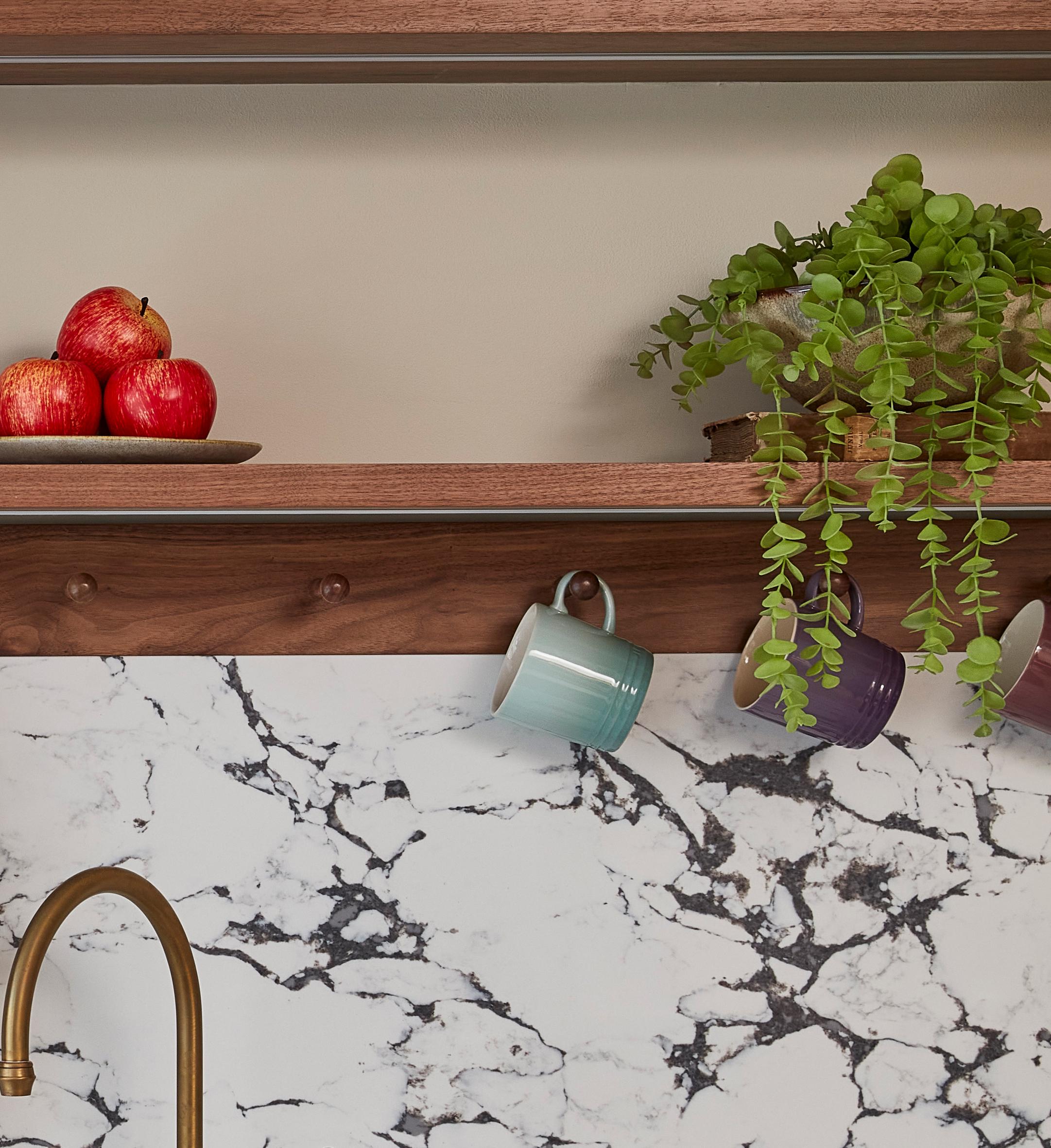 A kitchen scene with a marble backsplash and brass faucet. Wooden shelves hold a plate of red apples and a potted plant with trailing greenery. Three colorful mugs hang from hooks beneath the shelves