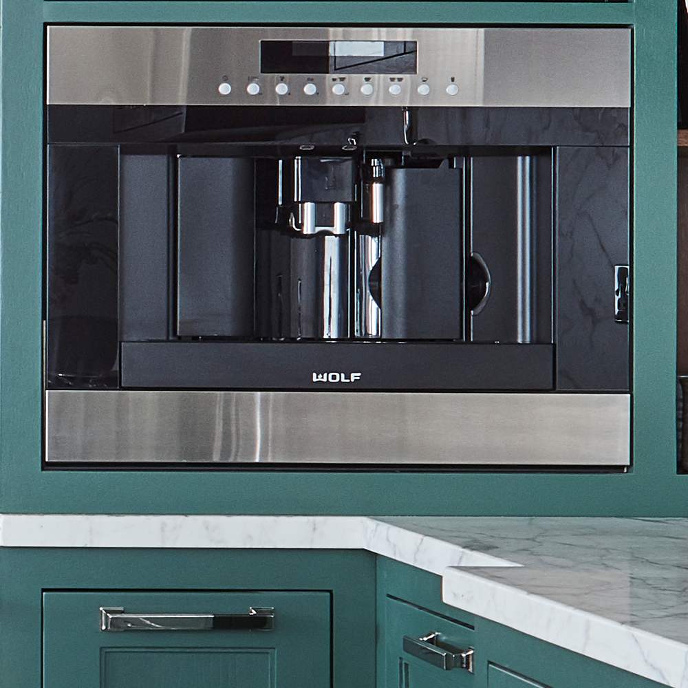 Built-in Wolf coffee maker with a stainless steel and black finish, installed in green cabinetry above a white marble countertop in a modern kitchen.