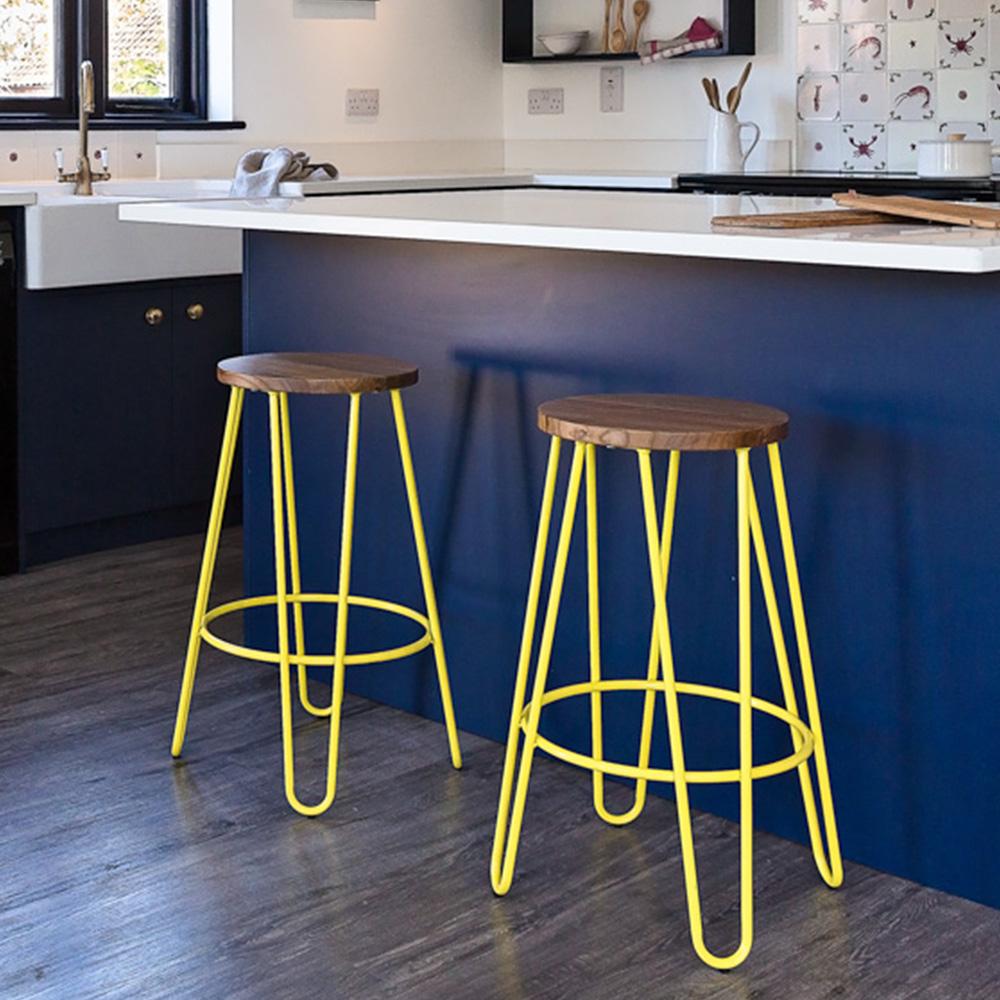 Two bar stools with yellow metal legs and wooden seats are placed in front of a blue kitchen island with a white countertop in a modern kitchen with dark wood flooring.