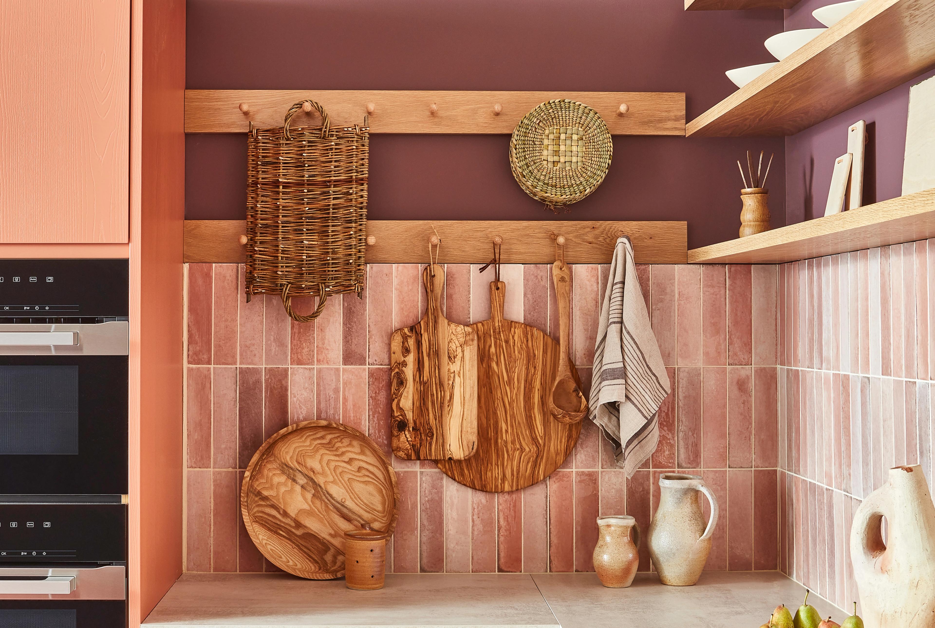 A warm-toned kitchen corner featuring wooden cutting boards hanging on a purple wall, a woven basket, and a patterned dish. Pink tiles line the backsplash, with shelves holding dishes and pottery accents. Neutral tones create a cozy atmosphere.