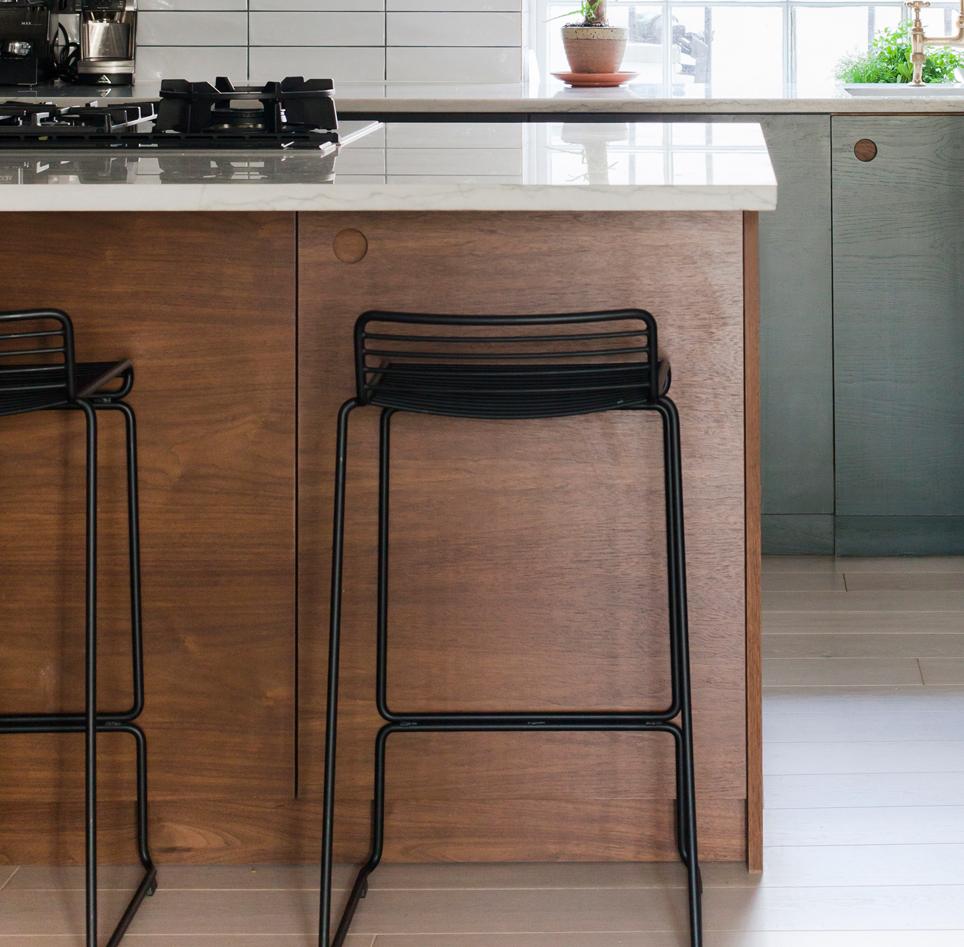 A modern kitchen features a wooden island with a black metal bar stool, a white countertop, a stove, green cabinets, and a small potted plant by a sunlit window.