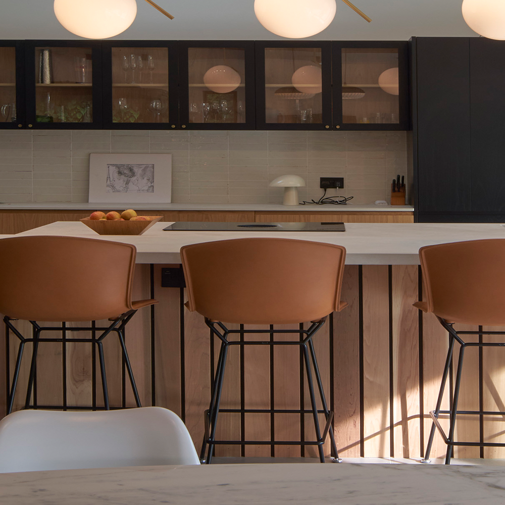 A modern kitchen with a wooden island and three brown barstools, glass-front cabinets, a bowl of apples, a small lamp, and minimalist decor under soft round pendant lights