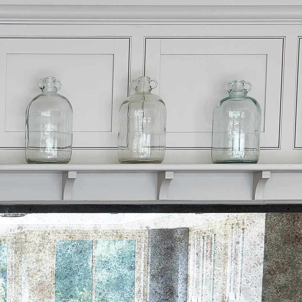 Three clear glass jugs are displayed on a white shelf against a white paneled wall, with a reflective surface and window light visible in the lower part of the image.