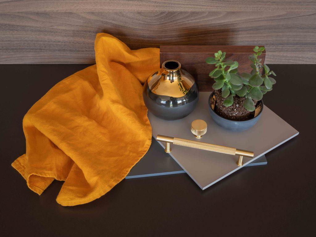 Interior design mood board flatlay featuring grey finish samples, knurled brass handles, a small metallic ceramic vase, a potted jade plant, and a draped mustard yellow linen cloth against a wood grain background.