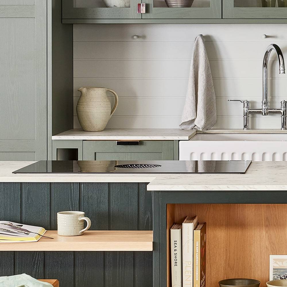 Modern kitchen with green cabinets, white countertops, and a sleek black stove. A pitcher and cloth are on the counter near a silver faucet. Below, a wooden shelf holds a mug and stacked books, adding warmth to the space.
