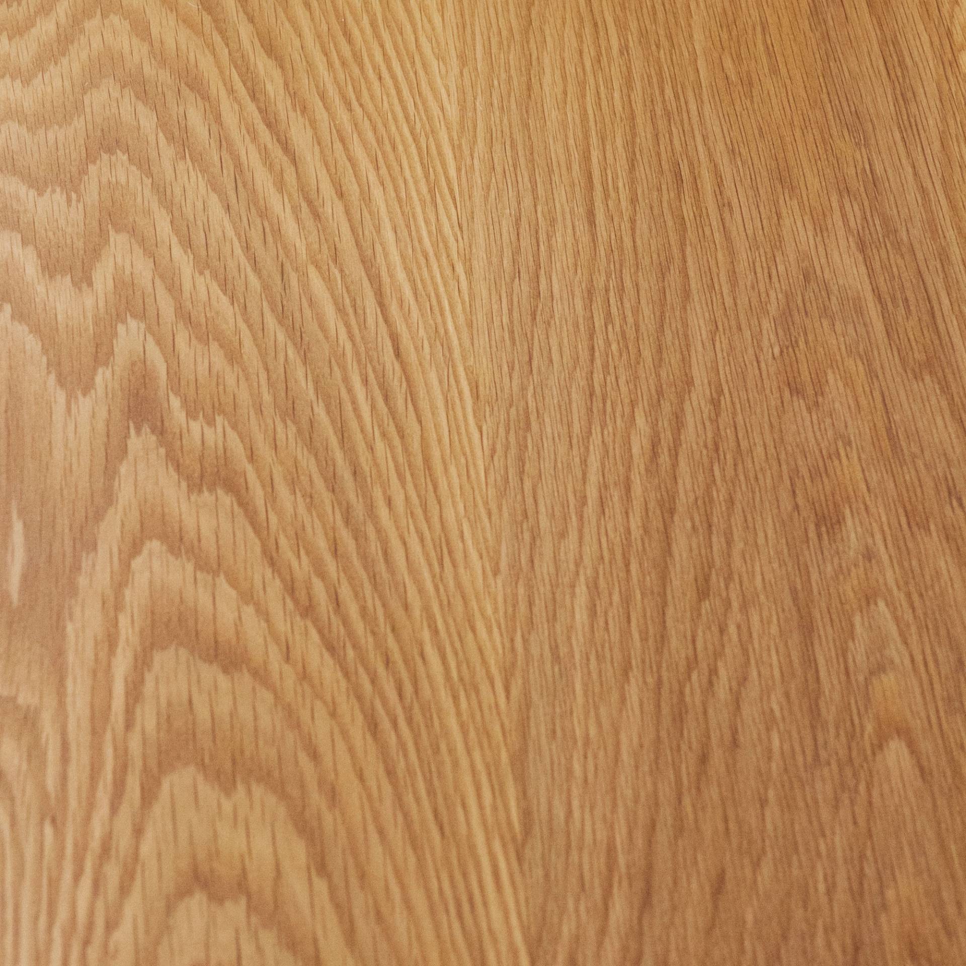 A close-up of a wooden surface showing the grain pattern. The wood features a light to medium brown hue with subtle variations and visible texture, highlighting the natural lines and striations typical of wood.