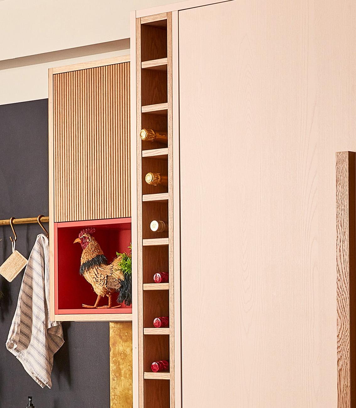 A modern kitchen cabinet with vertical storage slots holding wine bottles. A decorative rooster figure is displayed in a cubby with a red interior. A black wall with a gold rod holding a striped towel is partially visible.