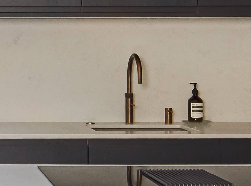 Modern kitchen sink with a sleek brass faucet, a built-in soap dispenser, and a bottle of liquid soap on a light countertop against a plain, light-colored backsplash.