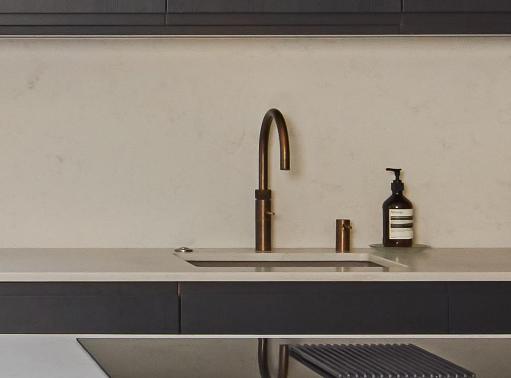 Modern kitchen sink with a sleek brass faucet, a built-in soap dispenser, and a bottle of liquid soap on a light countertop against a plain, light-colored backsplash.