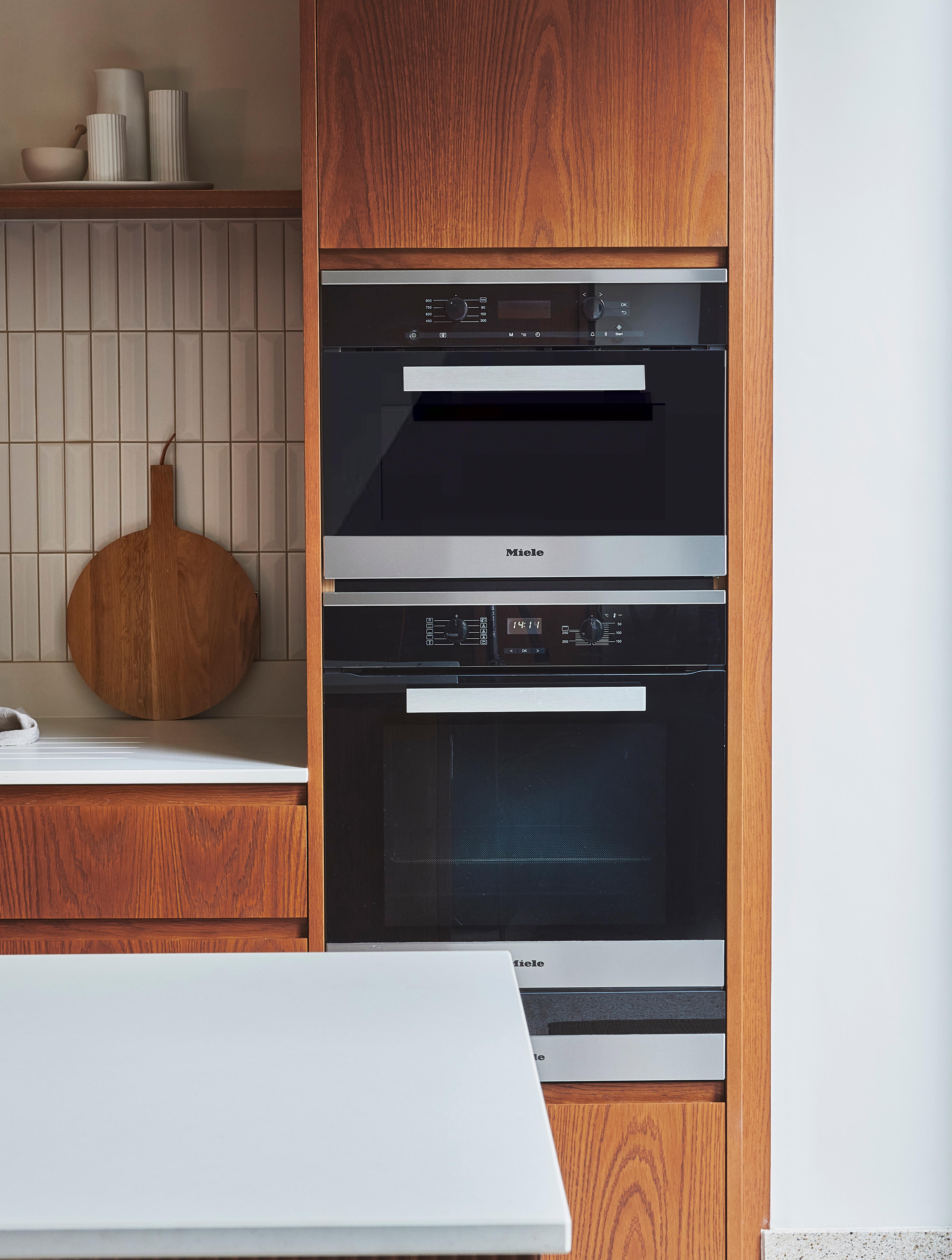 A modern kitchen with built-in wooden cabinets featuring a stacked oven and microwave. The countertop is light-colored, and theres a wooden cutting board on a side shelf against a tiled backsplash.