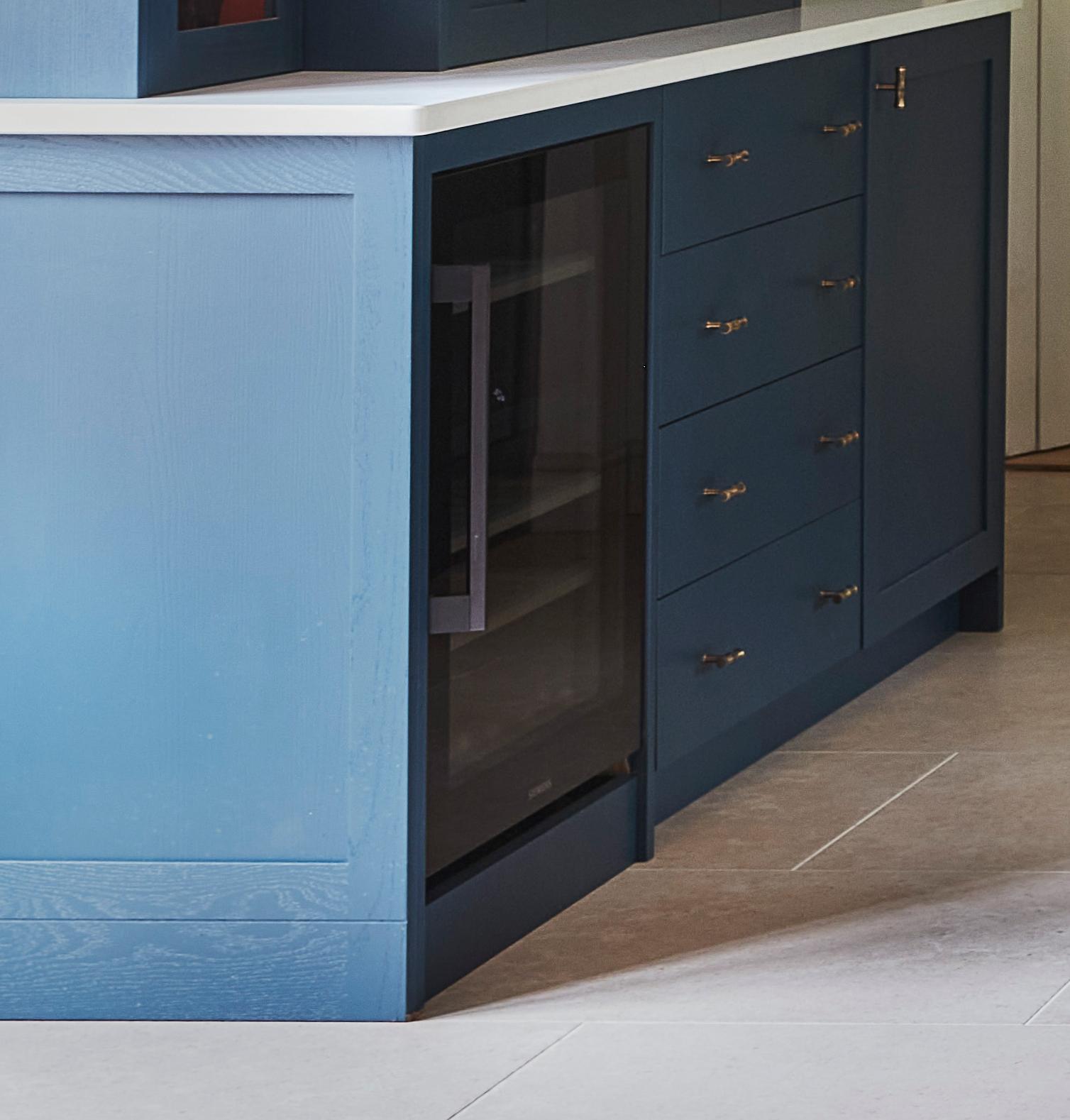 A blue kitchen island with a built-in glass-front mini fridge, several drawers with metal handles, and a white countertop on a light-colored tiled floor.