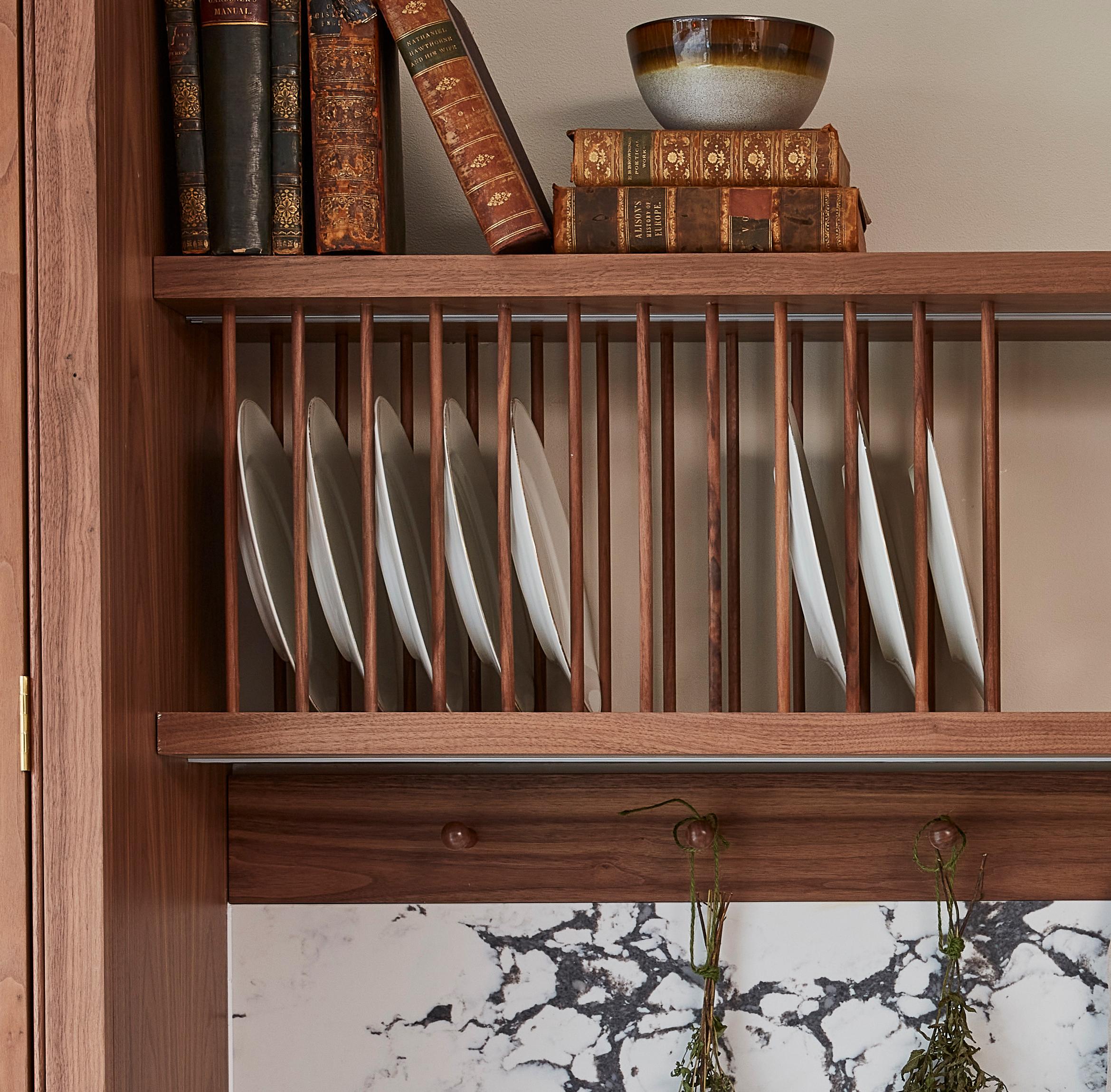 Wooden kitchen shelf with vertical slats holding white plates. Decorative ceramic bowls and old books are placed on top. Below, a marble backsplash features two sprigs of herbs hanging on hooks.