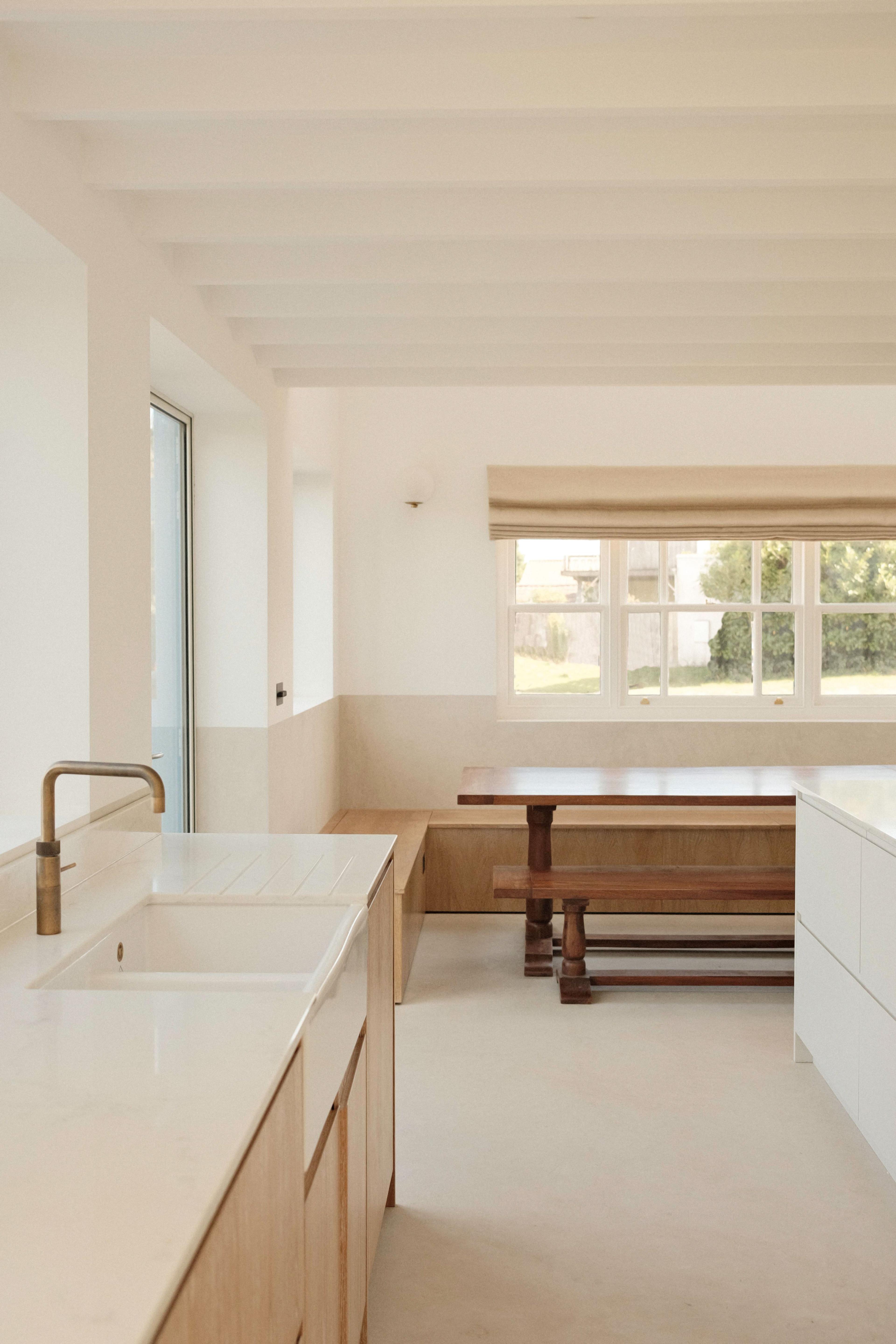 A bright, minimalist kitchen with white countertops, light wood cabinets, a built-in sink, large windows, and a wooden dining table with benches. Sunlight fills the space, highlighting its clean and modern design.
