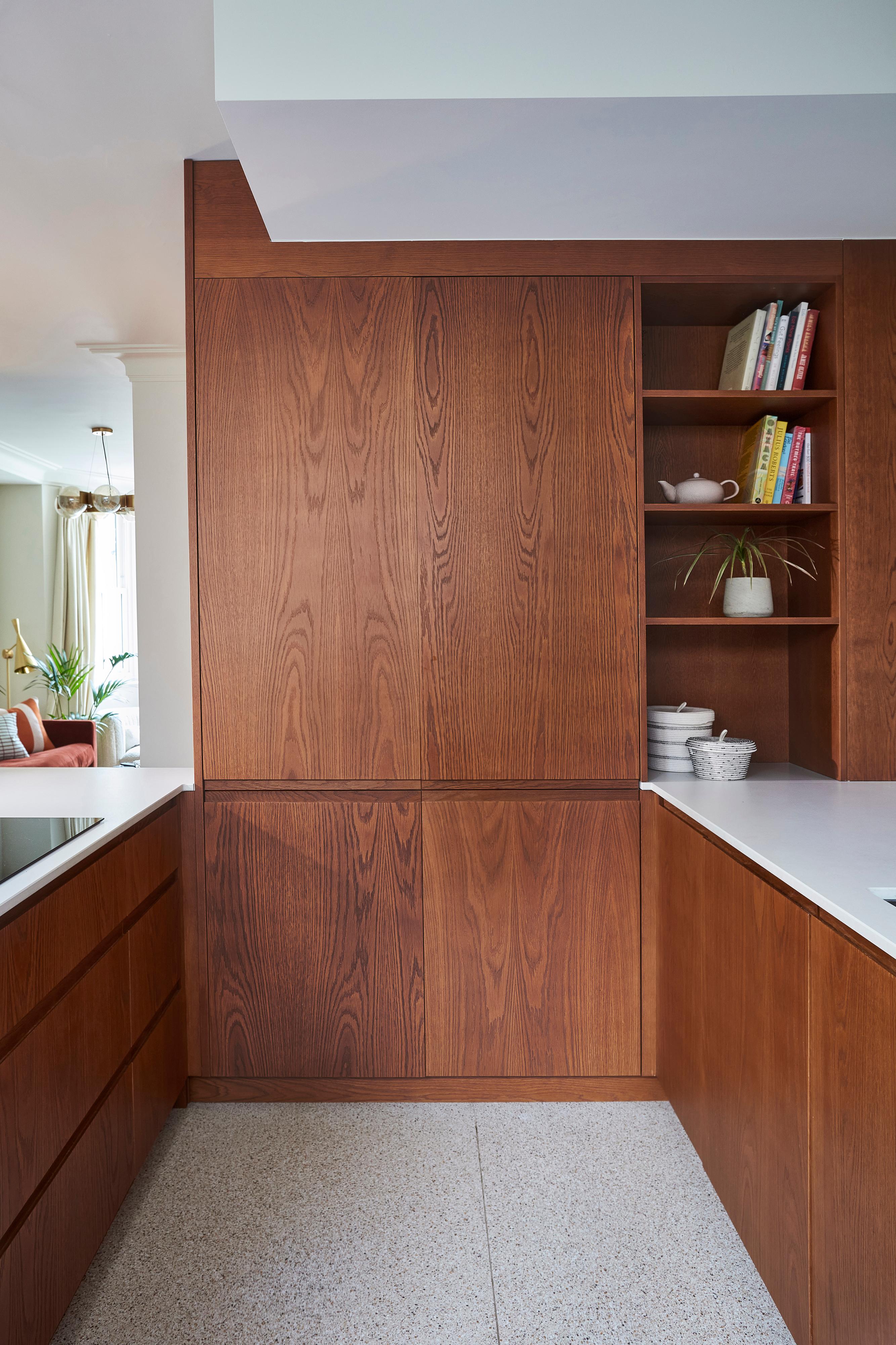 Modern kitchen with sleek wooden cabinets and white countertops. Open shelving displays books and a plant. Natural light streams in through a window, creating a warm, inviting atmosphere.