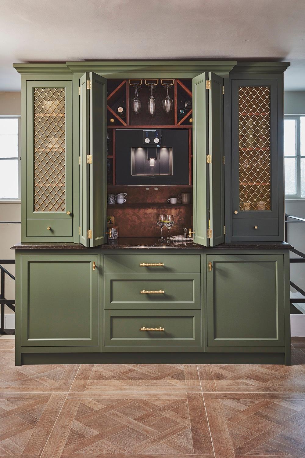An olive green drinks cabinet featuring folding doors that open to reveal a built-in coffee machine, wine racks, stemware, and brass accents over parquet wood flooring.