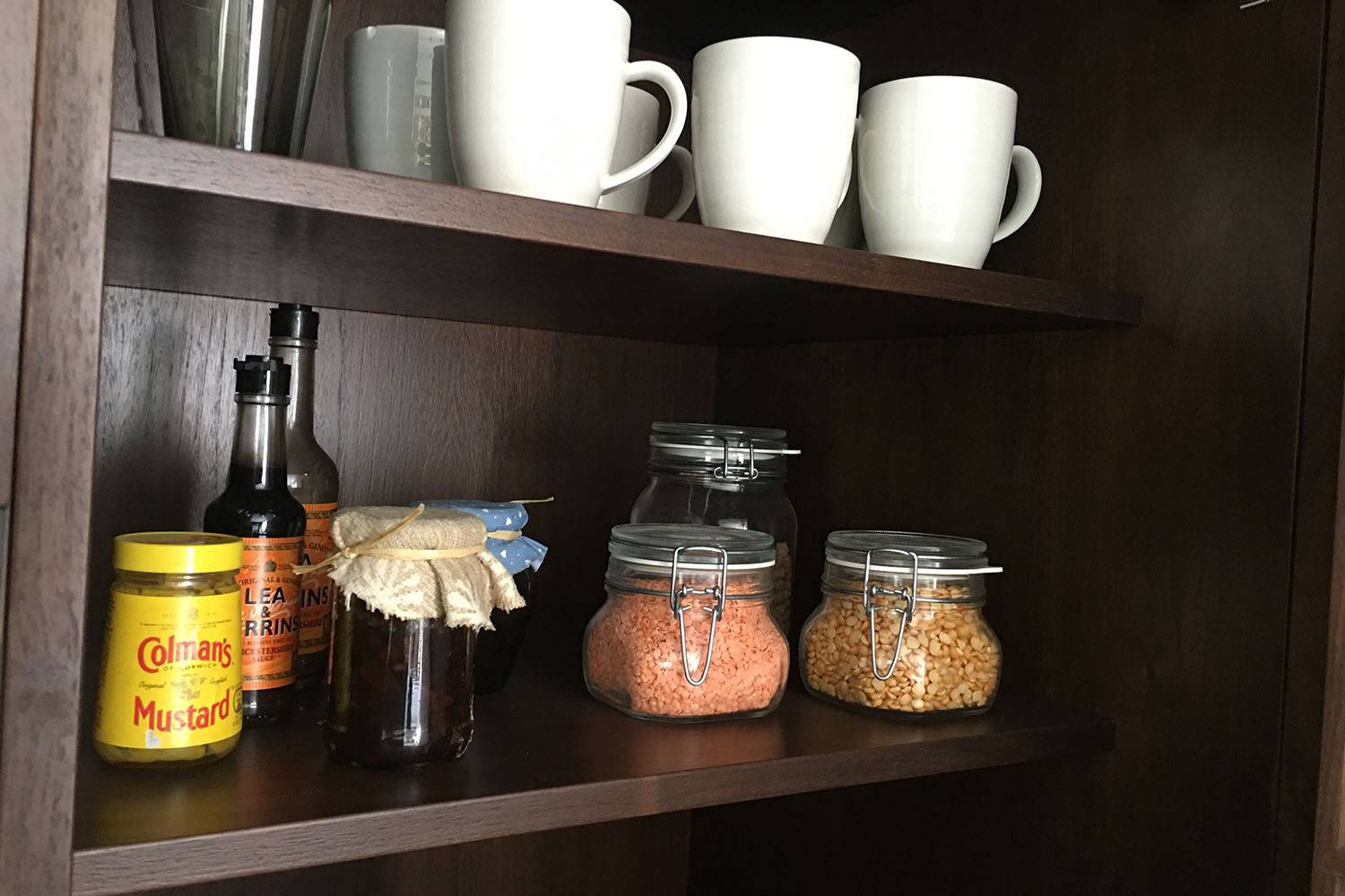 A wooden pantry shelf with white mugs on top. Below are bottles of Worcestershire sauce and a jar of English mustard. Two glass jars contain different grains or legumes. Another small jar is wrapped with a cloth cover.