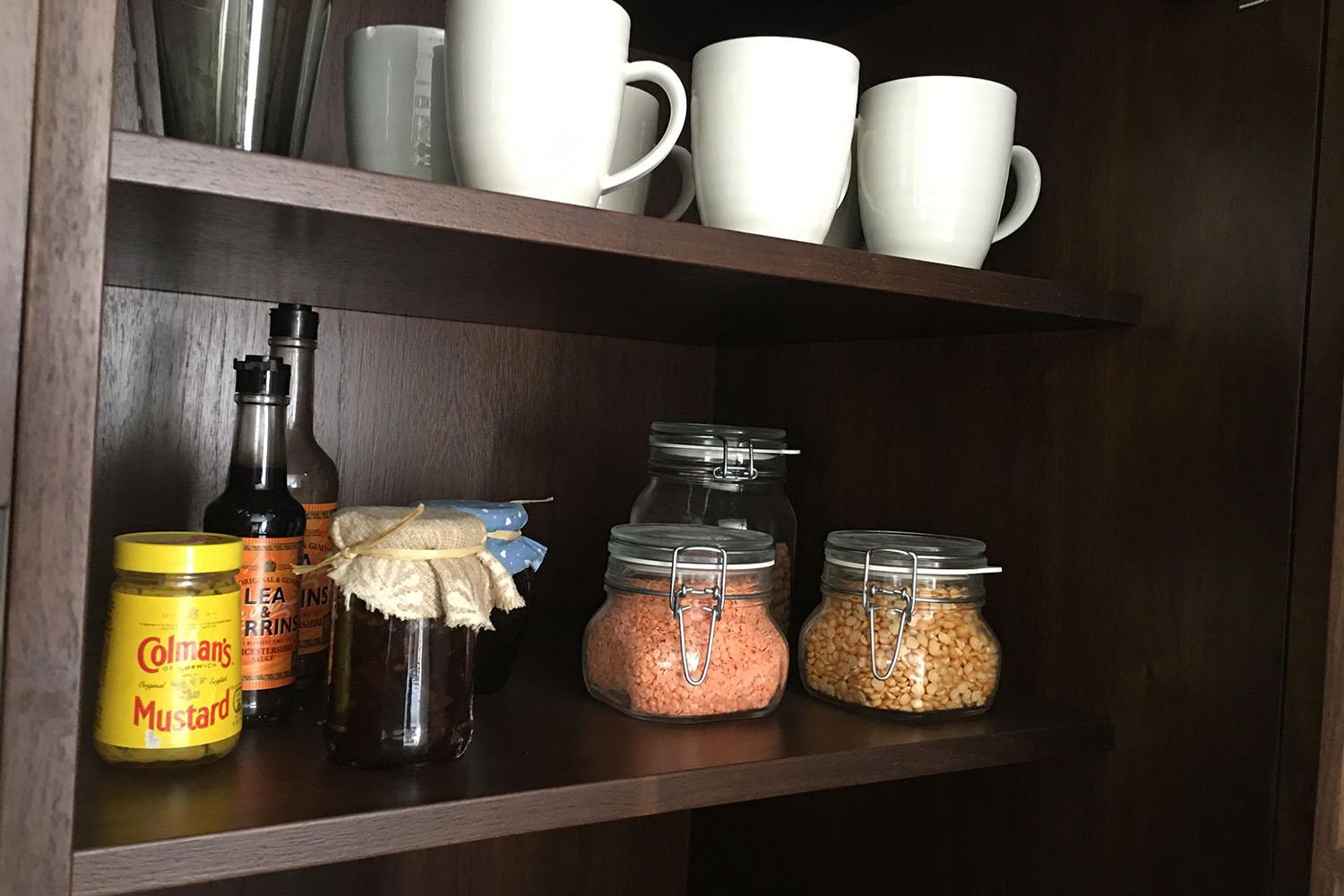 A wooden pantry shelf with white mugs on top. Below are bottles of Worcestershire sauce and a jar of English mustard. Two glass jars contain different grains or legumes. Another small jar is wrapped with a cloth cover.