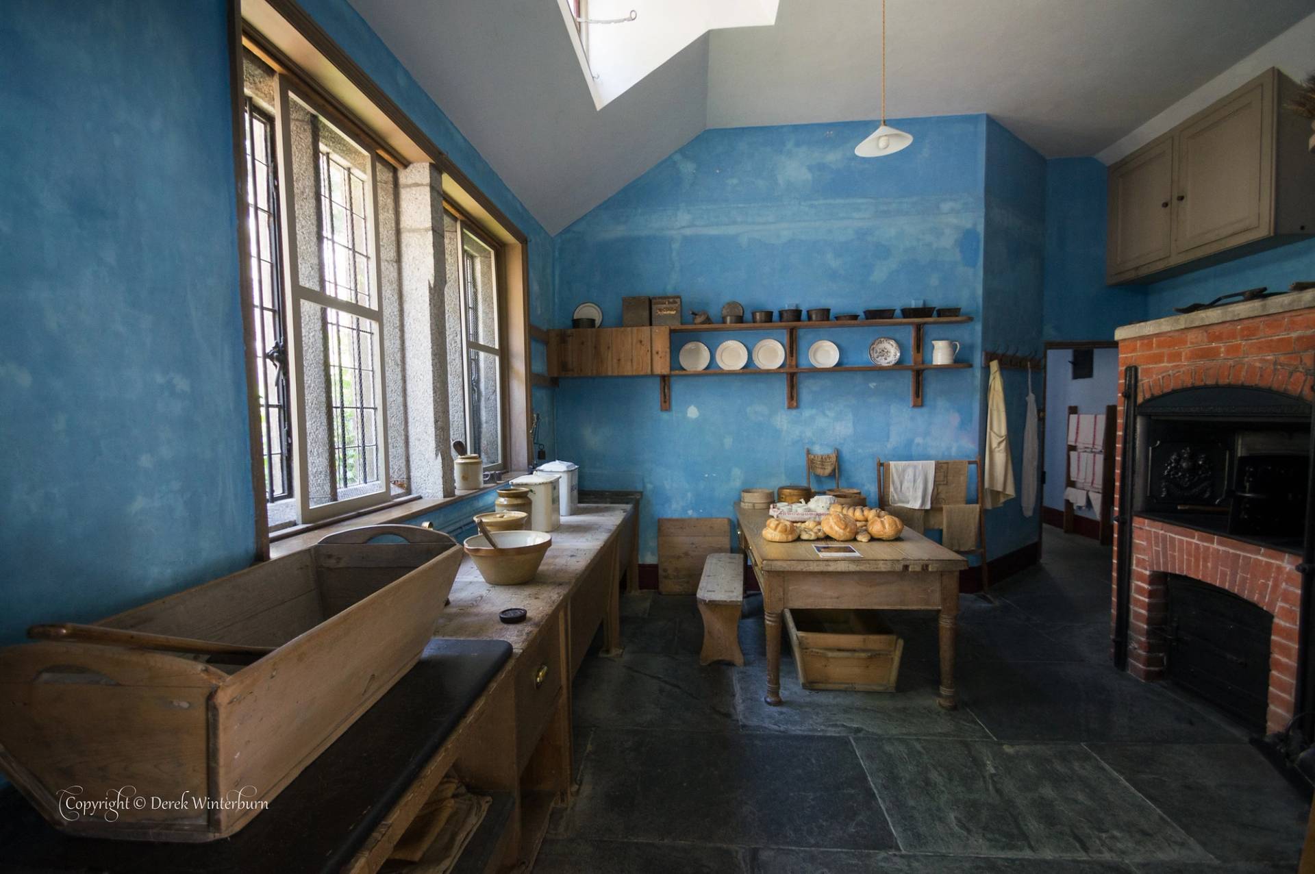 A historic kitchen with textured blue walls, a brick hearth, and flagstone flooring, featuring a central wooden prep table with bread loaves, wall-mounted display plates, and a long countertop with large wooden troughs and mixing bowls by deep-set windows.