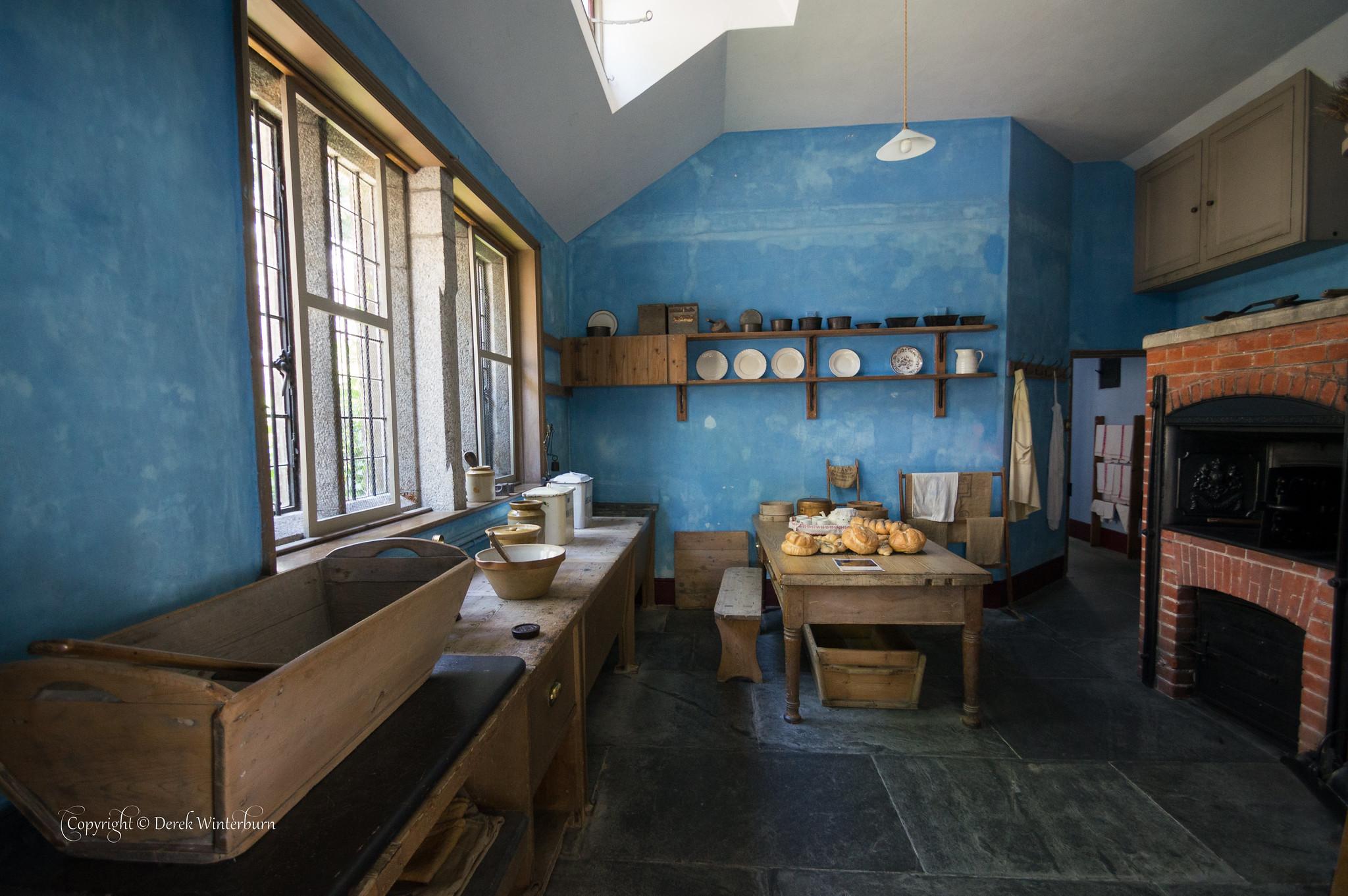 A historic kitchen with textured blue walls, a brick hearth, and flagstone flooring, featuring a central wooden prep table with bread loaves, wall-mounted display plates, and a long countertop with large wooden troughs and mixing bowls by deep-set windows.