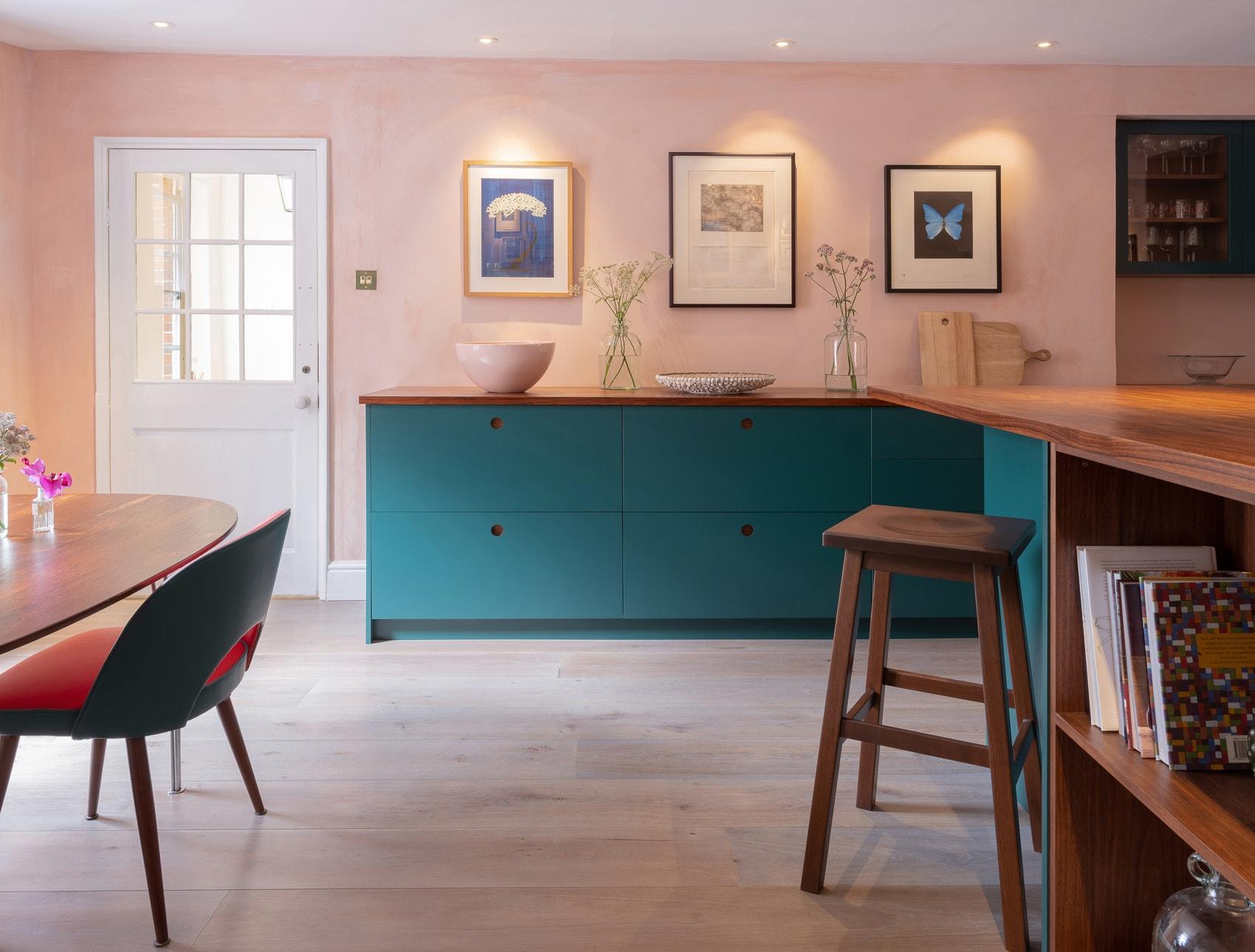 A cozy kitchen with light pink walls and wooden floors. A wooden counter and bar stool are in the foreground. A teal cabinet with art above, and a small white bowl with flowers, adorn the back wall. A door with glass panels is to the left.