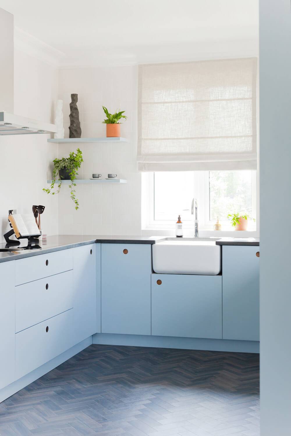 A modern kitchen with pastel blue cabinets, a white farmhouse sink, and a wooden zigzag-patterned floor. Two small shelves hold plants and decor. A large window with a light curtain brightens the room.