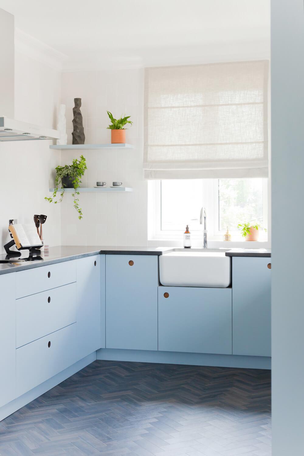 A modern kitchen with pastel blue cabinets, a white farmhouse sink, and a wooden zigzag-patterned floor. Two small shelves hold plants and decor. A large window with a light curtain brightens the room.