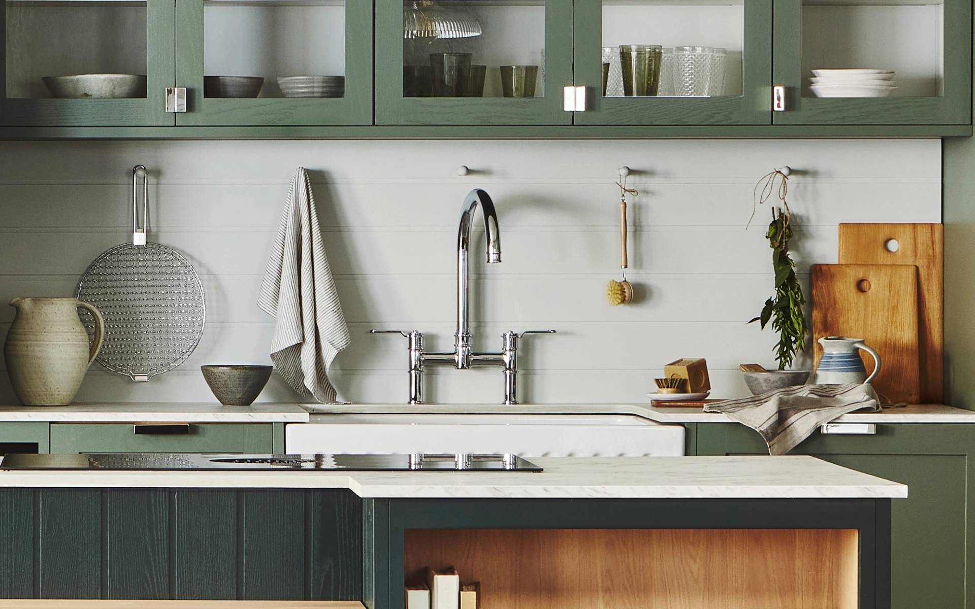A modern kitchen with green cabinetry, white walls, and a farmhouse sink. The countertop has a cutting board, pitcher, and two bowls. Shelves display glassware and dishes. A towel hangs by the sink, and a plant adds a touch of nature.