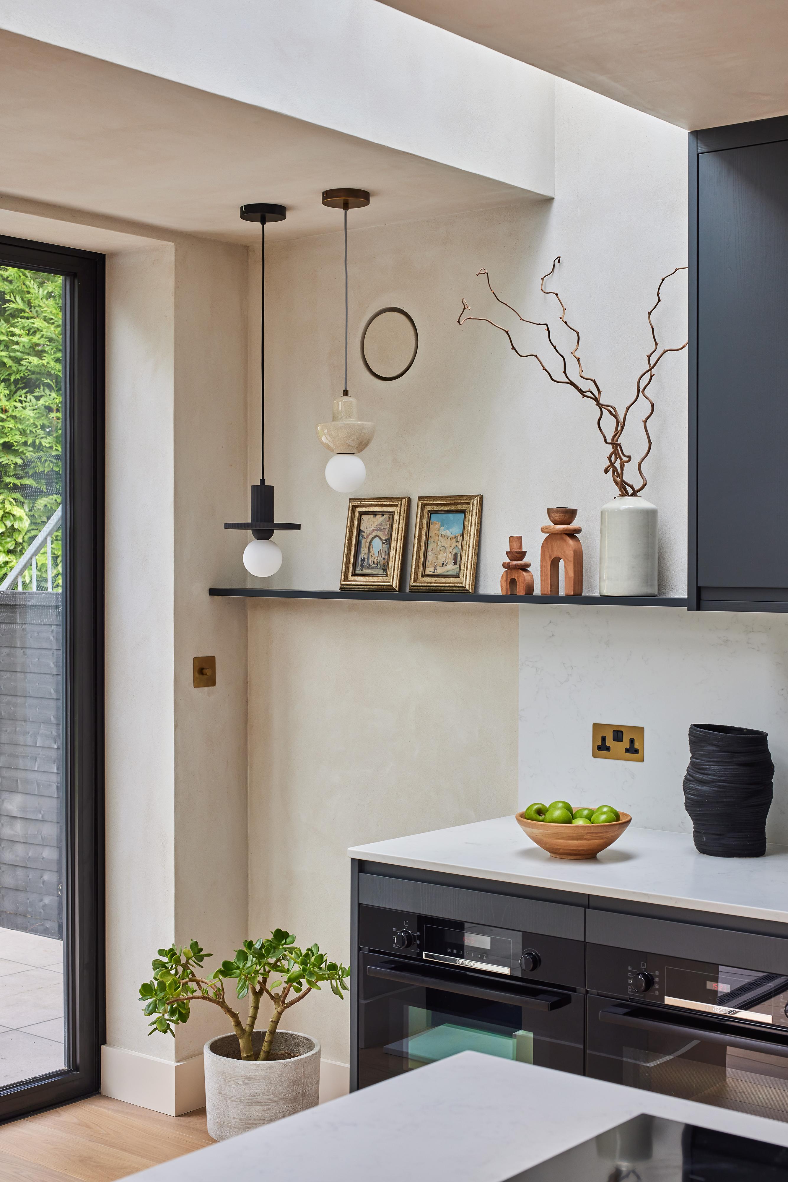 Modern kitchen with dark cabinets, a white countertop, and a potted plant on the floor. Shelf with framed pictures, vases, and decor. Two pendant lights hang from the ceiling; a bowl of green apples sits on the counter.