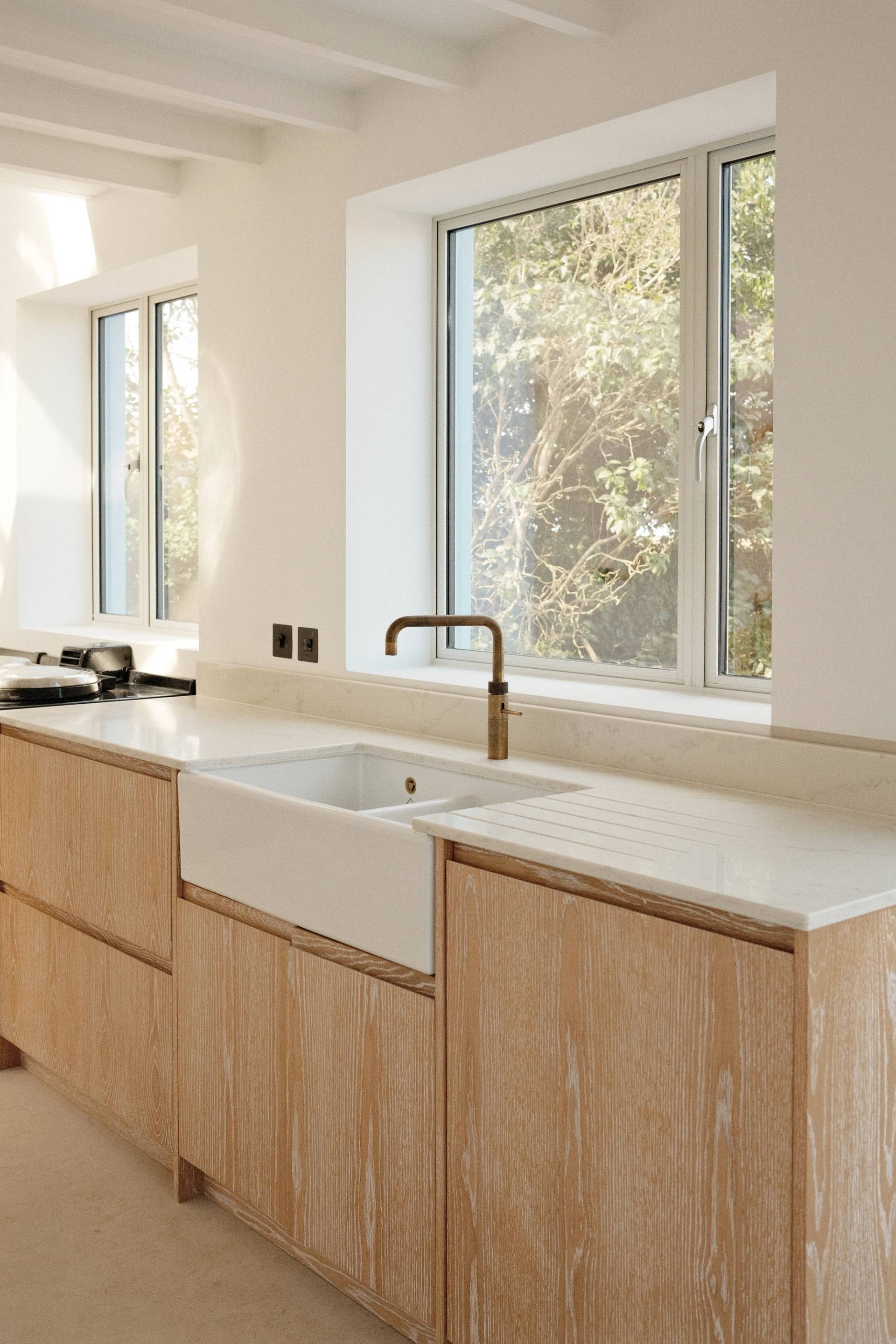 a bright, minimalist kitchen features light-colored wooden cabinets, a white marble countertop with an integrated farmhouse sink, and a brass faucet positioned in front of a large window overlooking green trees.