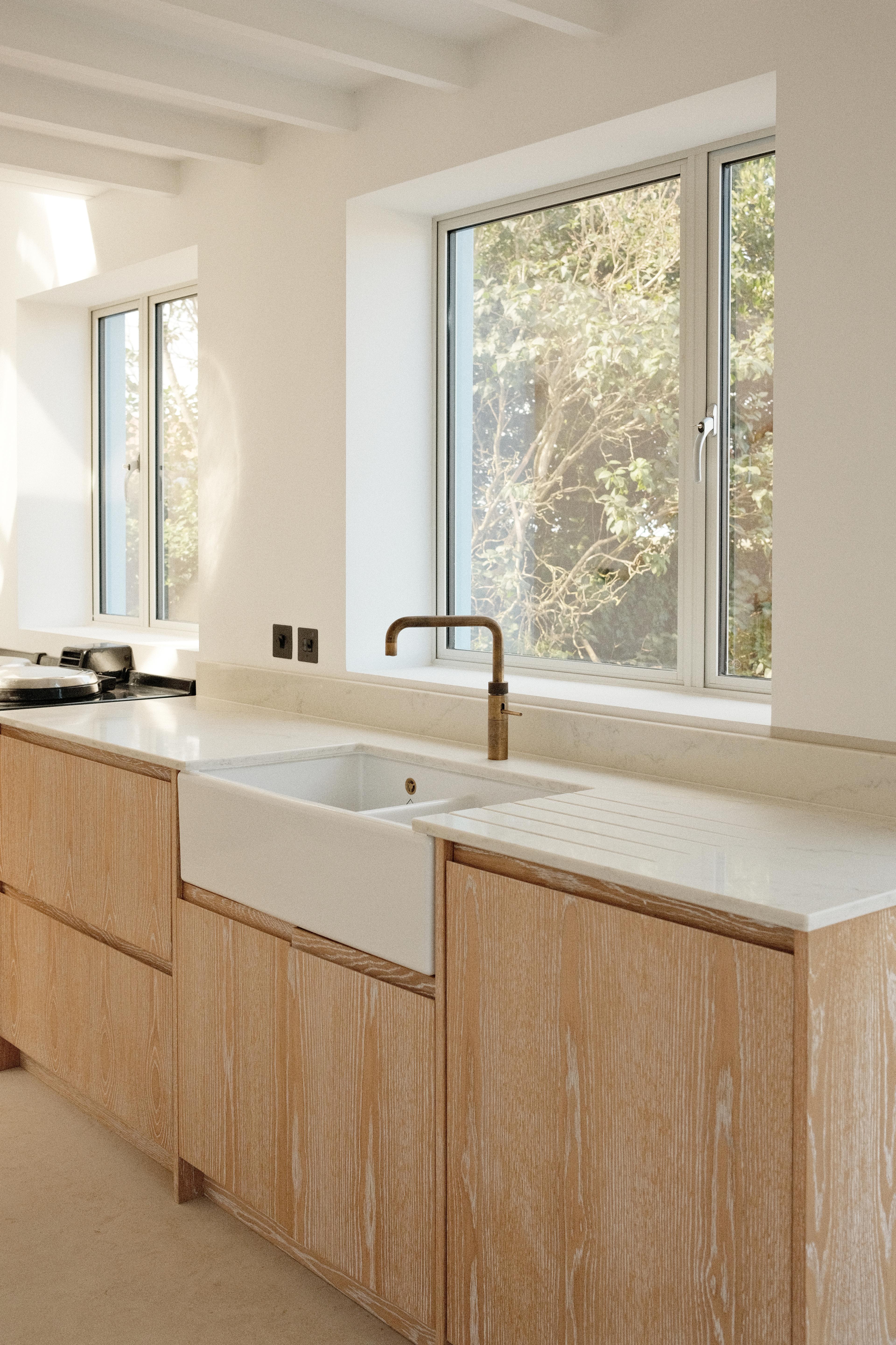 a bright, minimalist kitchen features light-colored wooden cabinets, a white marble countertop with an integrated farmhouse sink, and a brass faucet positioned in front of a large window overlooking green trees.