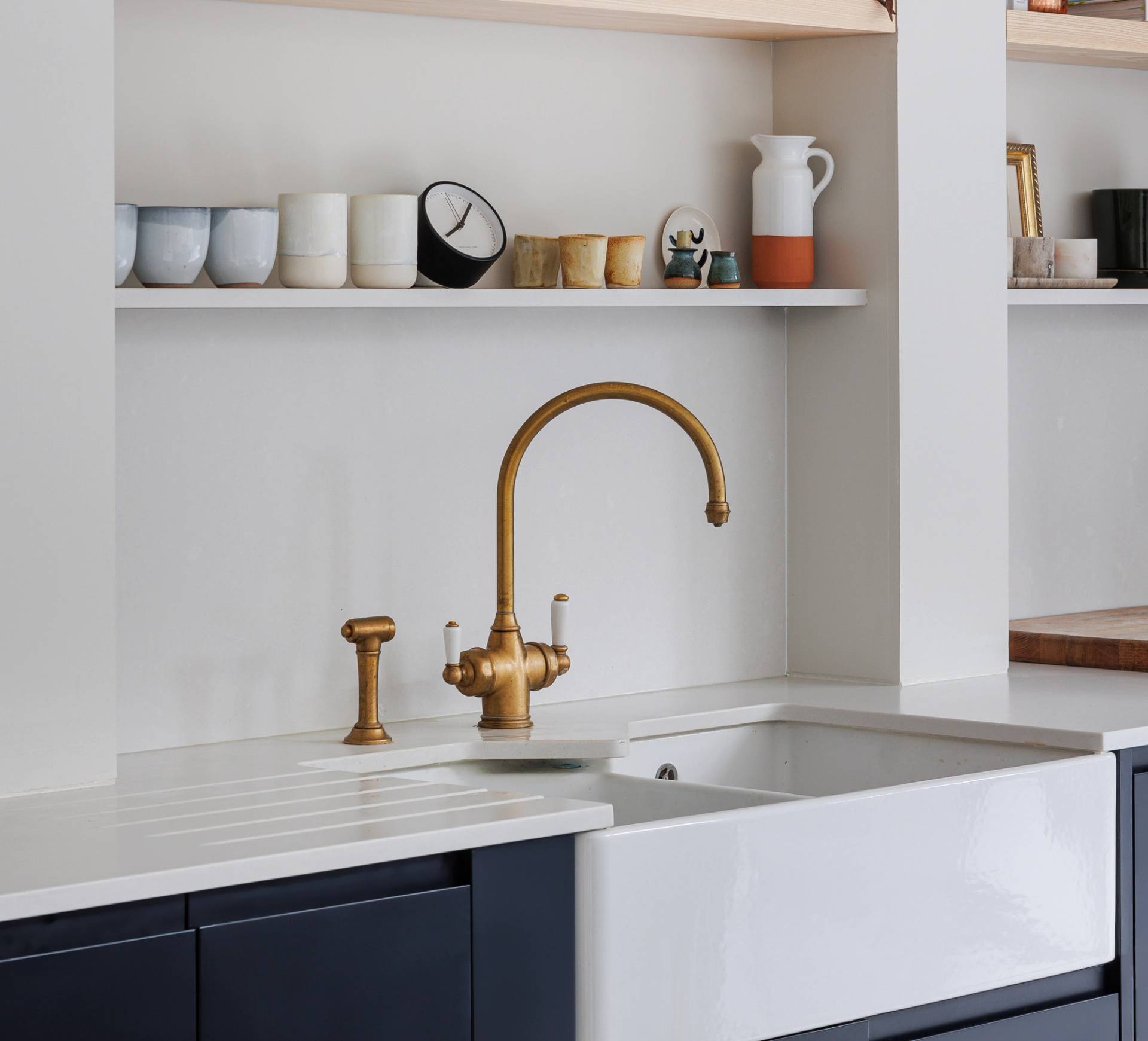 A modern kitchen sink with a gold faucet and soap dispenser sits below white shelves holding various cups, bowls, a clock, and decor items. The countertop and backsplash are white with dark lower cabinets.