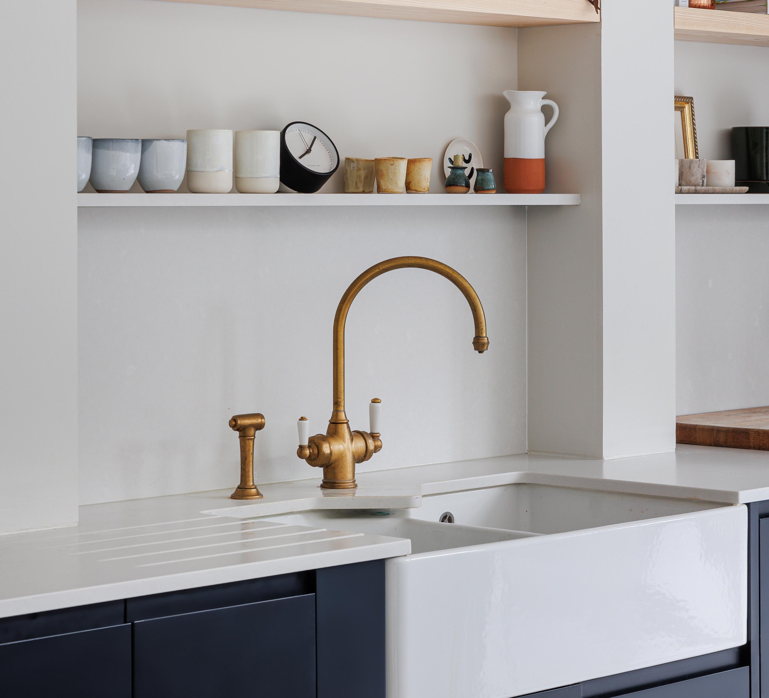 A modern kitchen sink with a gold faucet and soap dispenser sits below white shelves holding various cups, bowls, a clock, and decor items. The countertop and backsplash are white with dark lower cabinets.