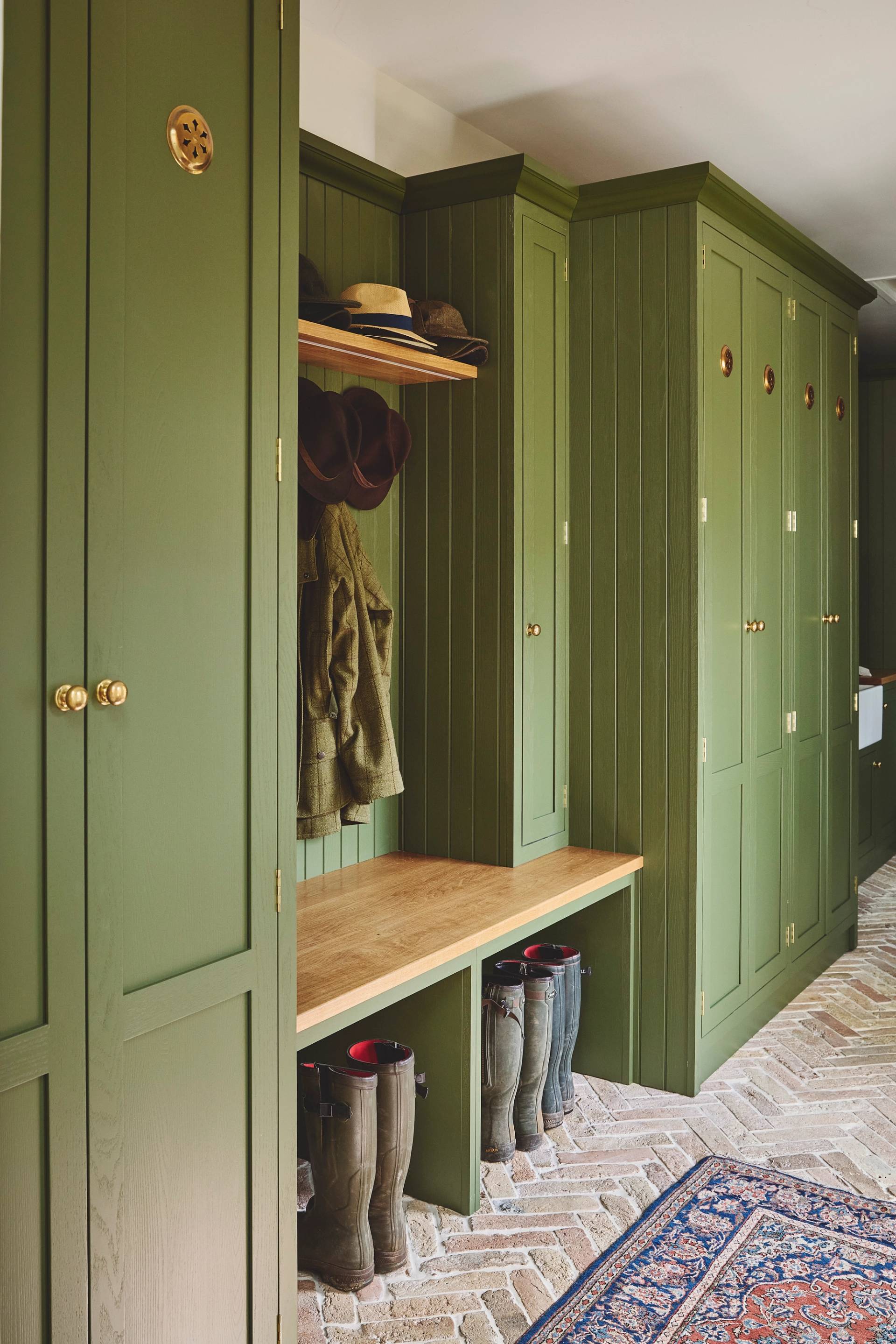 A mudroom with olive green paneled cabinets, brass hardware, a wooden bench, boots lined underneath, coats and hats on hooks, and a patterned rug on a herringbone brick floor.