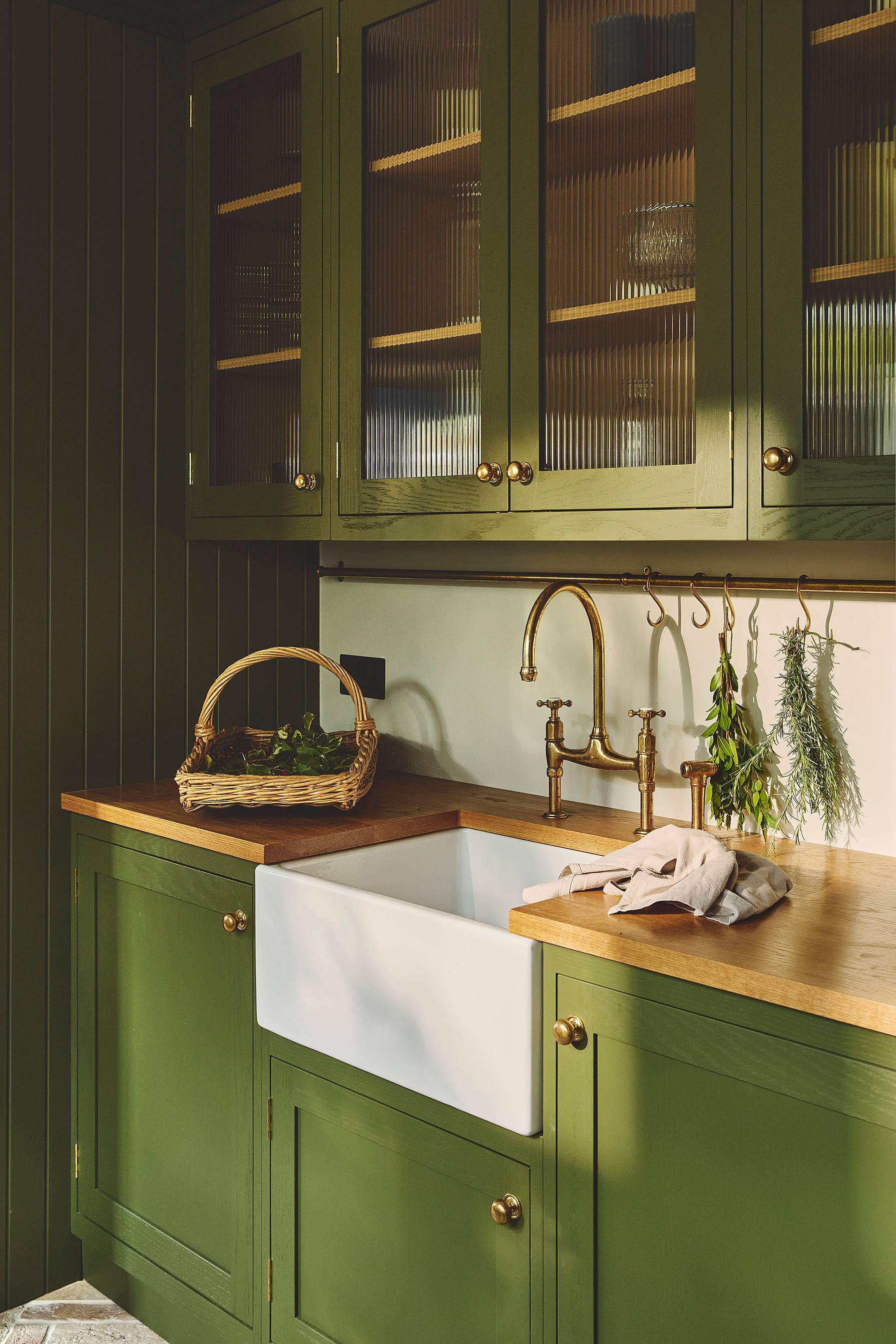 dark green shaker cabinetry with a butler sink in the centre. The tap is brass coloured. The worktop is oak and is covered in fresh herbs. There is a basket with more herbs in the background. There are wall cabinets with glazed glass fronts.