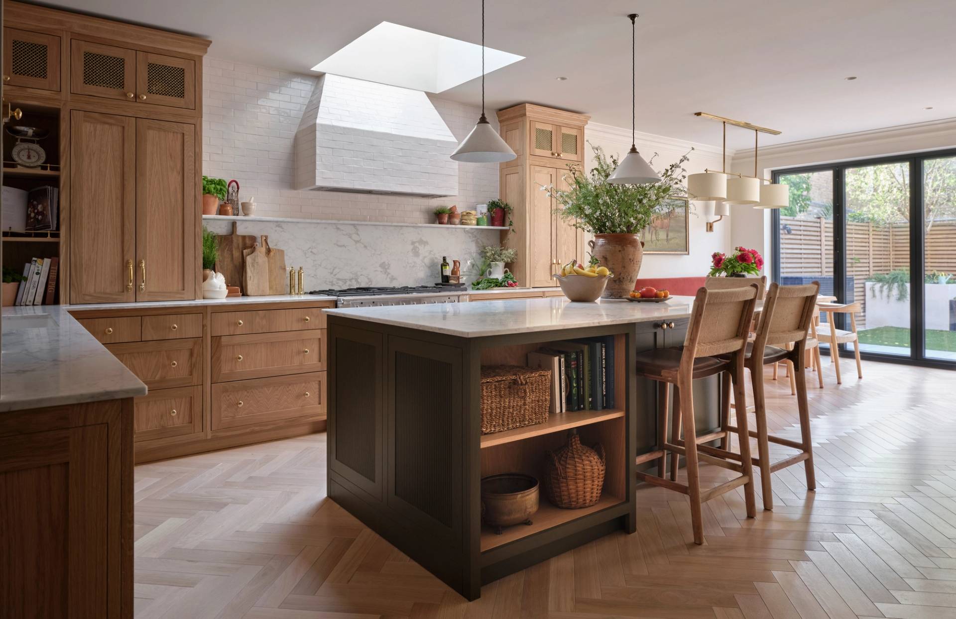 A spacious, modern kitchen with light wood cabinets, a large island with shelves, pendant lights, marble countertops, and large windows opening to a garden. There are plants and fruit bowls on the island and counters.