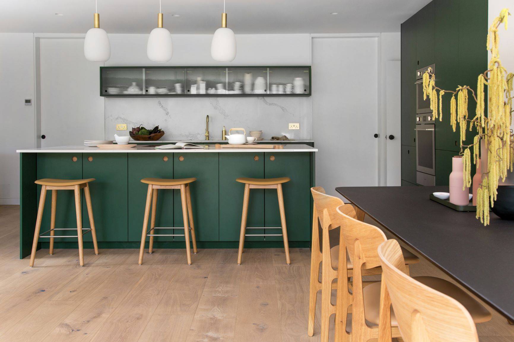 Modern kitchen with green cabinets and a large island featuring wooden stools. Hanging pendant lights above the island, and a dining table with wooden chairs. Sleek appliances and minimal decor add a contemporary feel. Light wood flooring throughout.