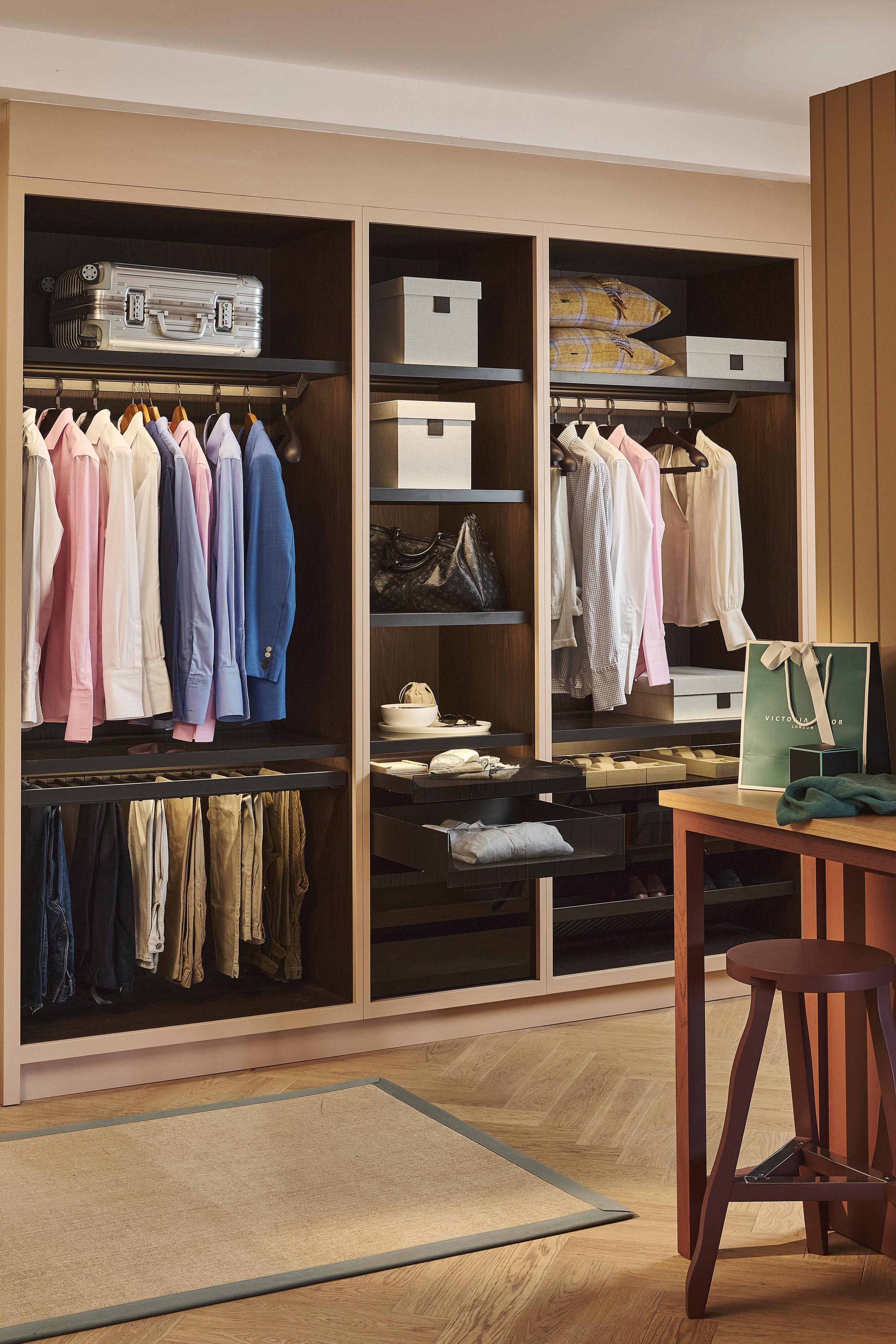 A neatly organized closet with shirts, jackets, and pants hanging, shelves with boxes and bags, folded clothes, and a suitcase. A wooden stool and shopping bag are visible in the foreground.